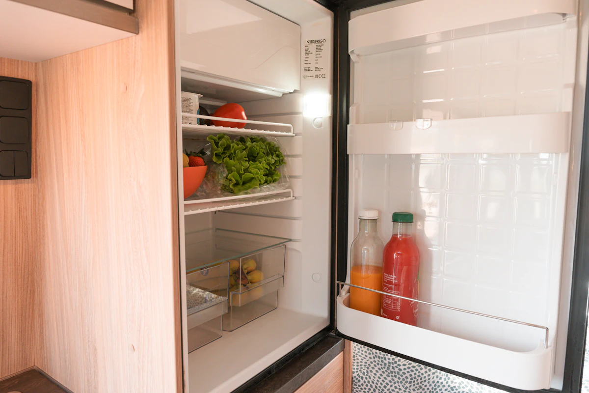 Open campervan fridge with fresh vegetables, fruits, and drinks stored neatly inside on shelves and fridge door.