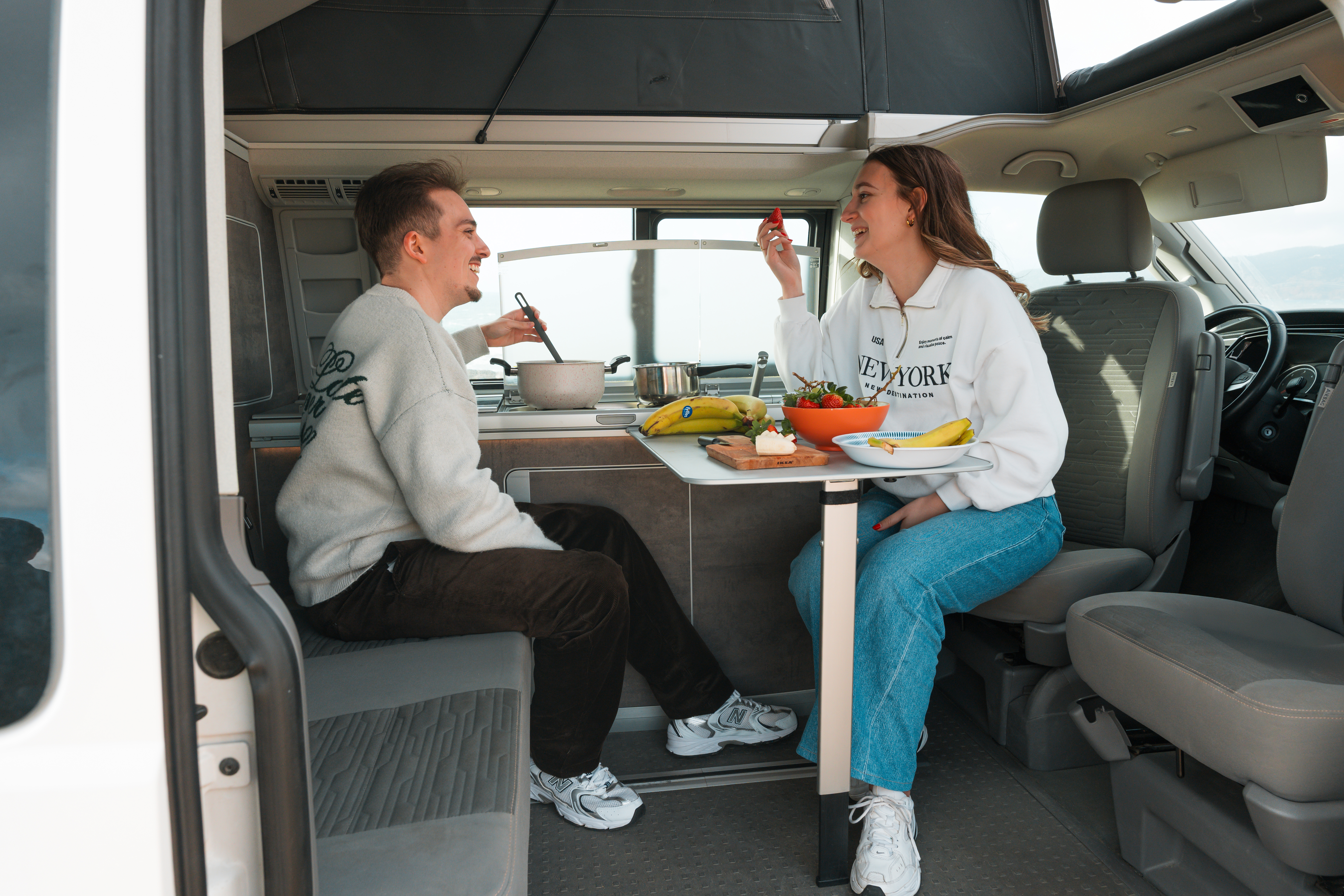 Two people sitting inside a camper van, enjoying a meal together at a table with food and drinks.