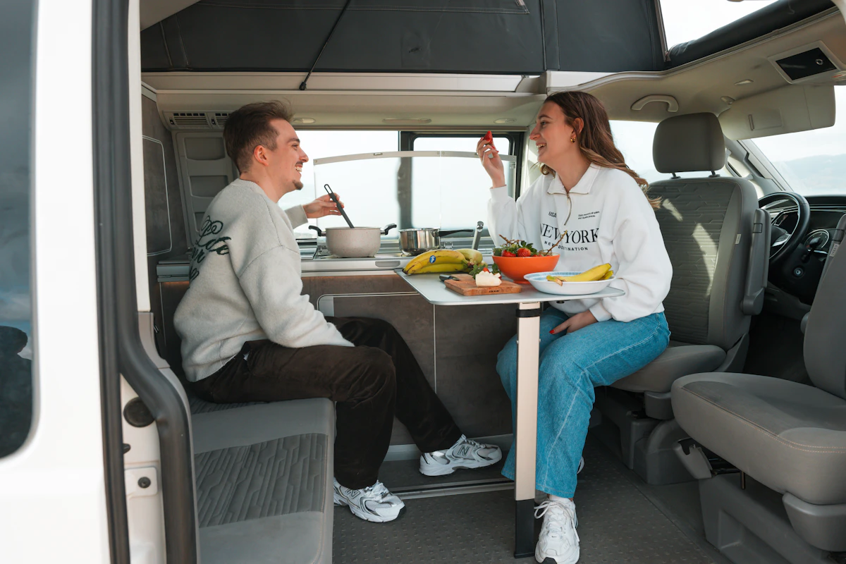Two people sitting inside a camper van, enjoying a meal together at a table with food and drinks.