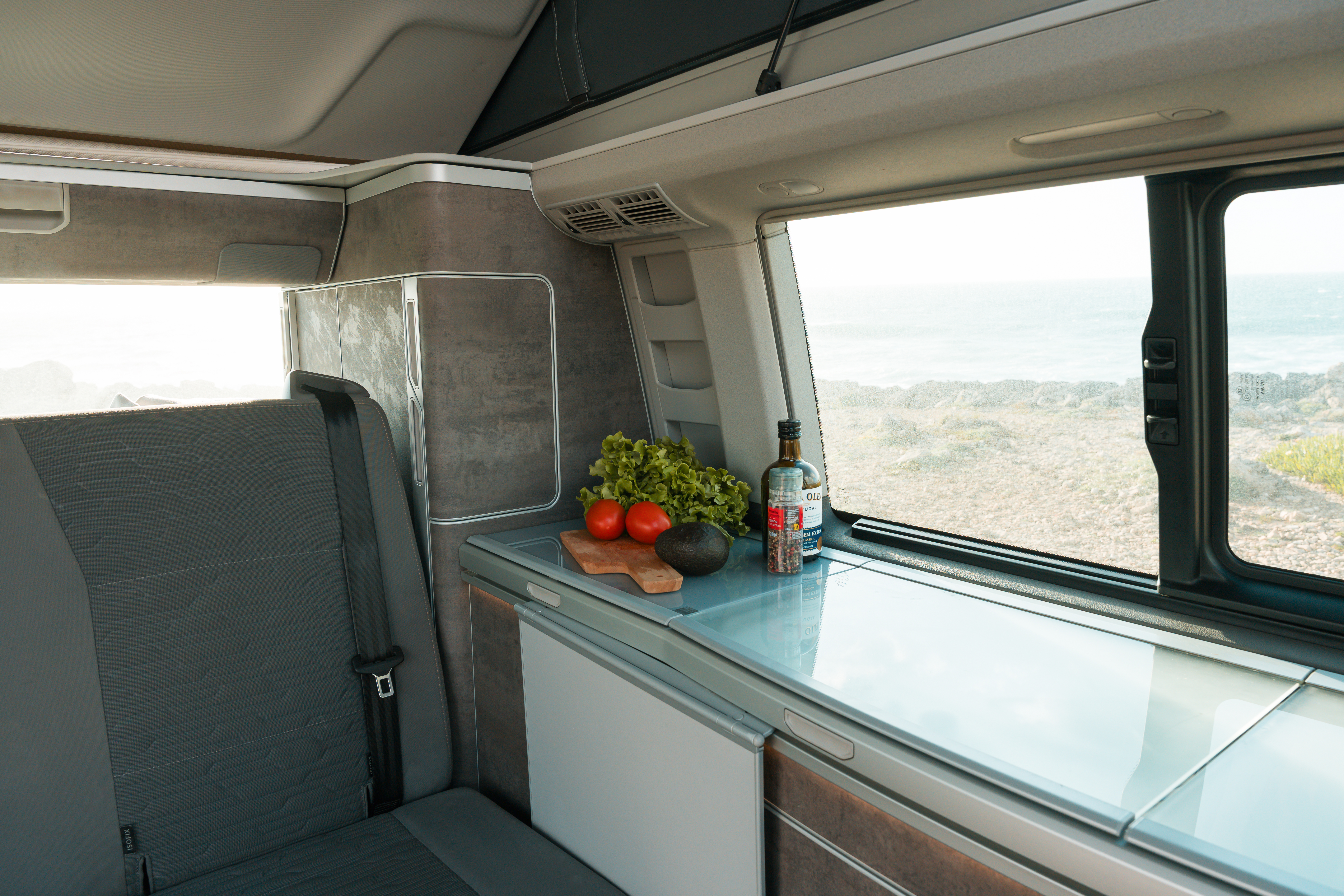 Interior of a campervan kitchen with vegetables, fruit, and a bottle on a counter by a window.