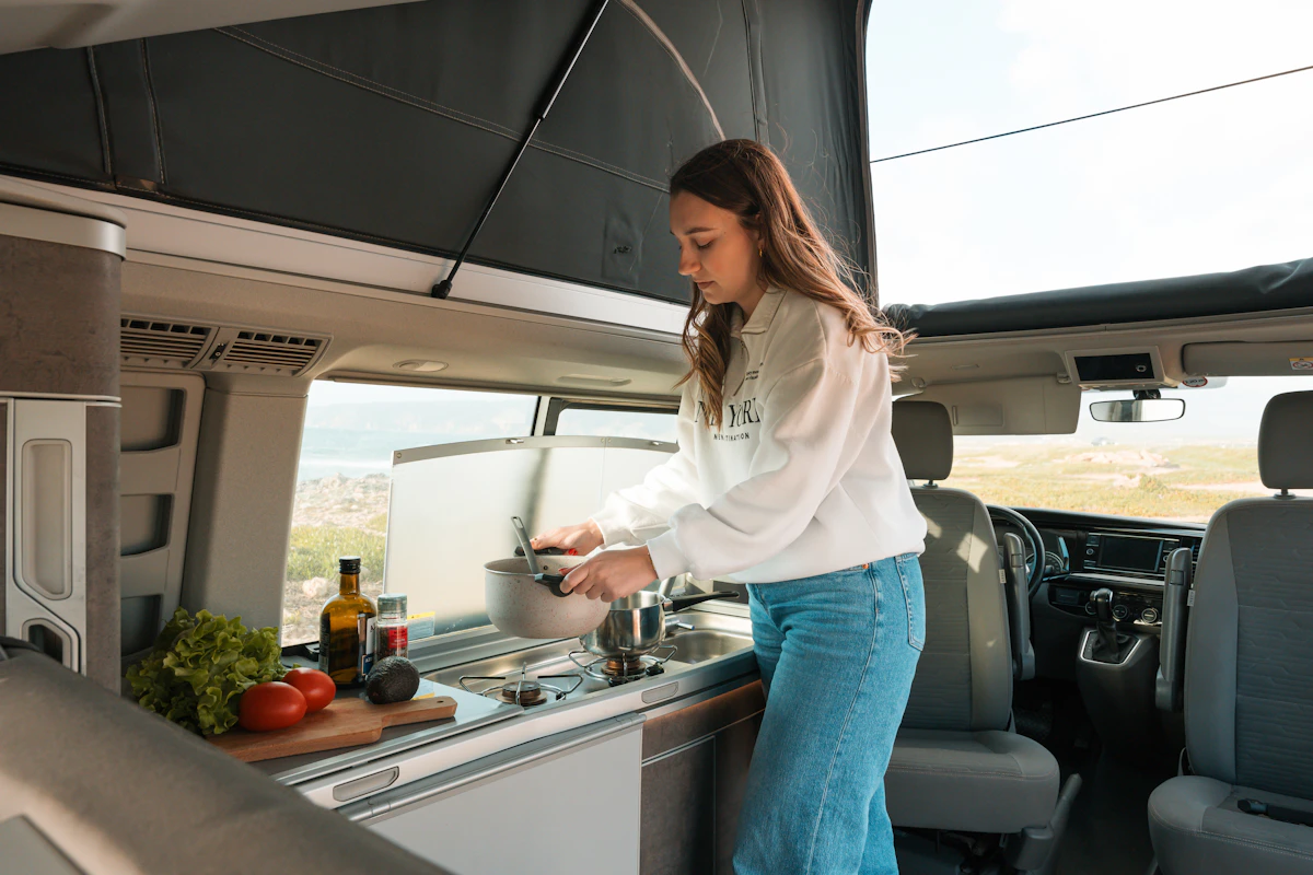 Woman cooking in a campervan kitchen with fresh vegetables and condiments on the countertop, interior view.