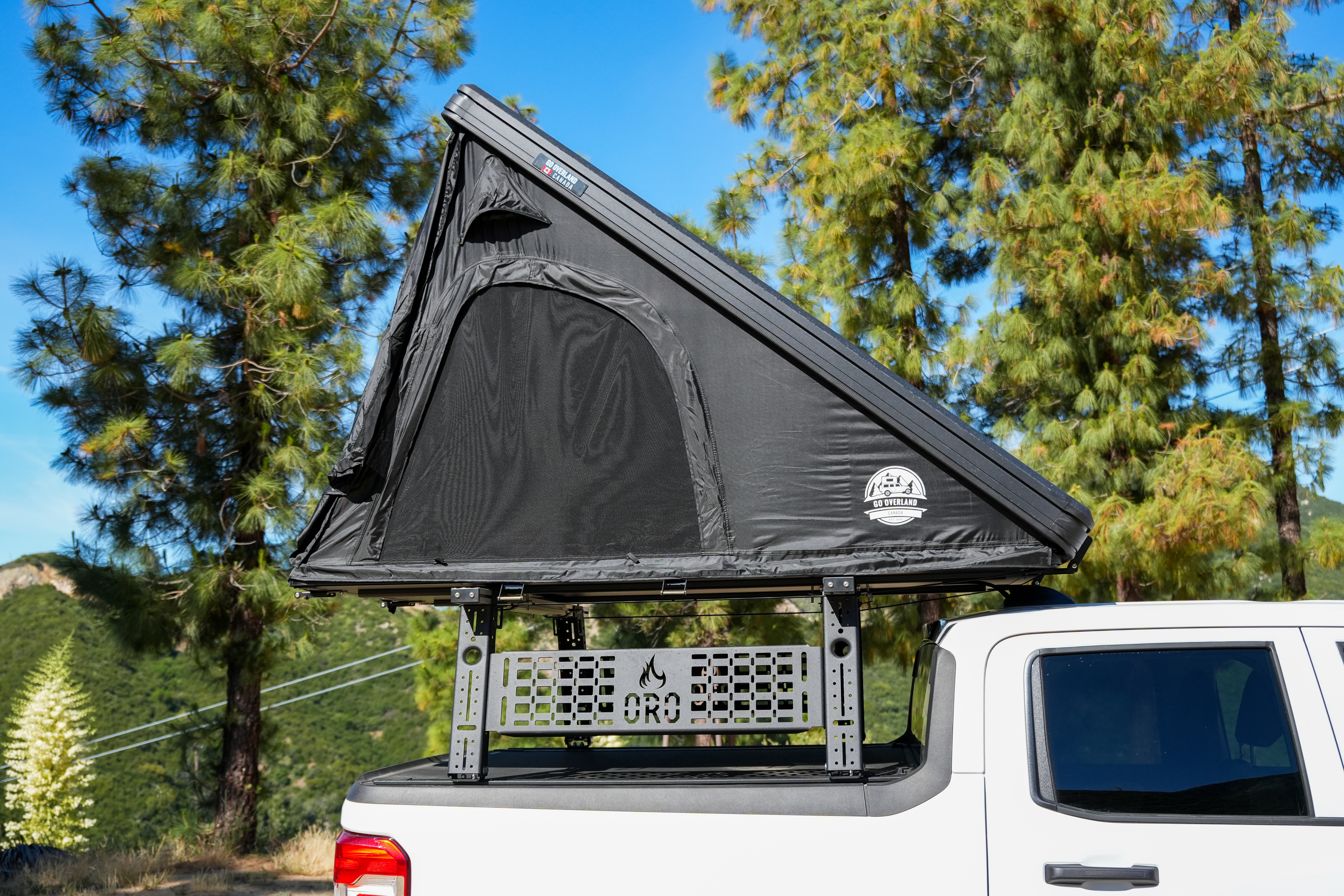 Foldable black rooftop tent mounted on a white pickup truck, surrounded by pine trees in a natural outdoor setting.