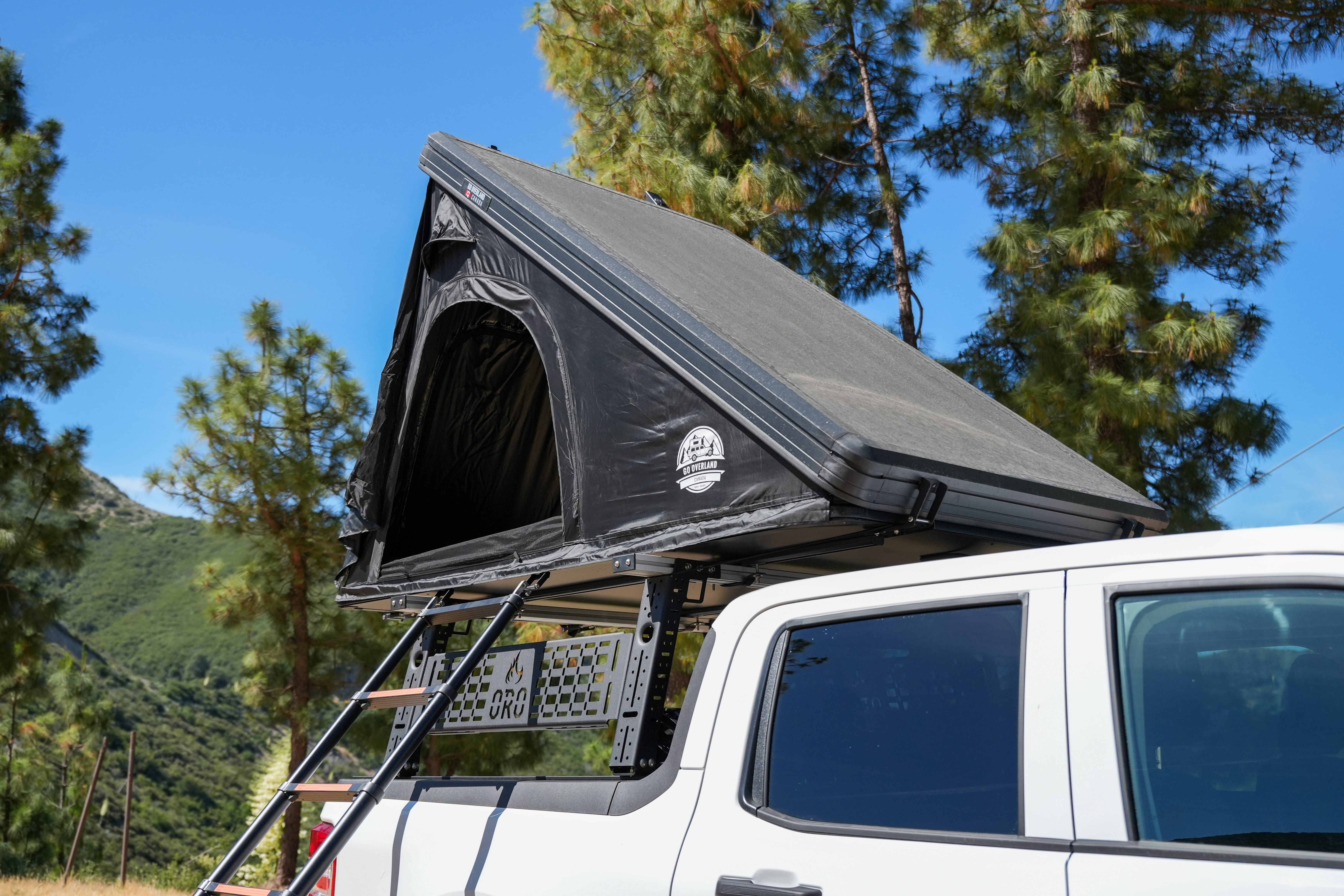 Rooftop tent mounted on a white camper van with a ladder, set in a scenic outdoor location with trees.