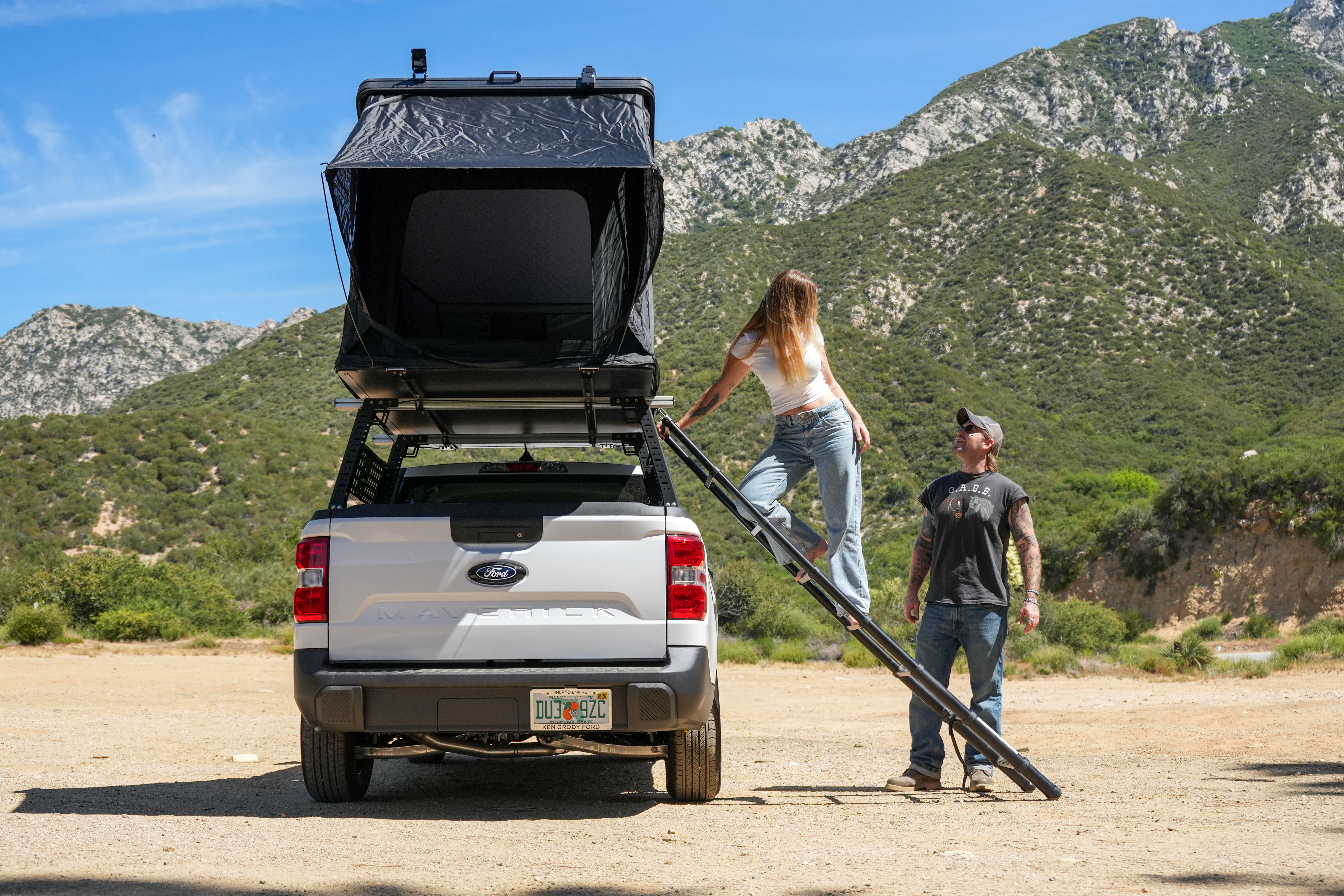 Pickup truck with rooftop tent, woman climbing ladder, man standing nearby, mountains and blue sky in the background.