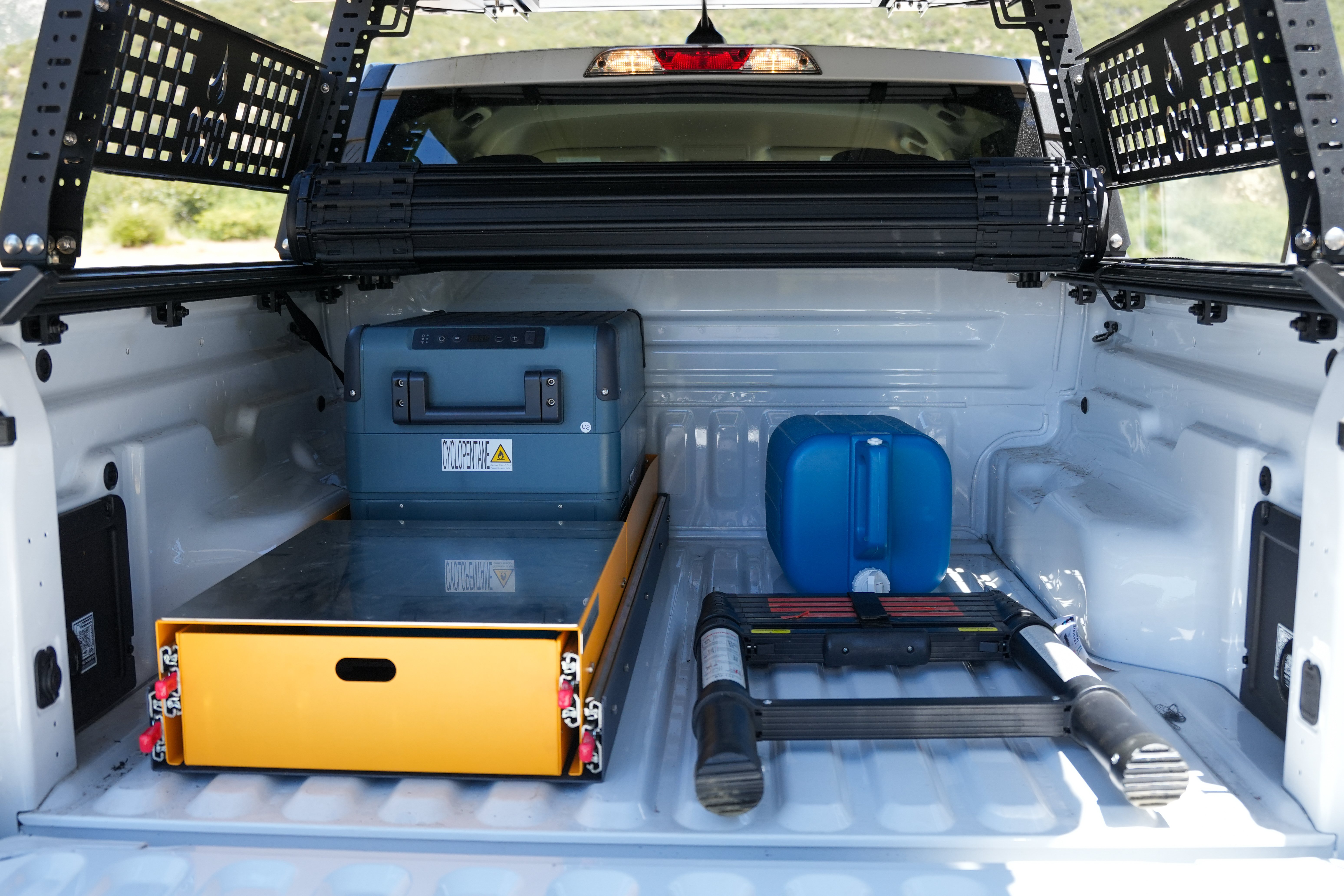 Truck bed equipped with storage drawer, portable fridge, blue water container, and foldable ladder, ready for camping.