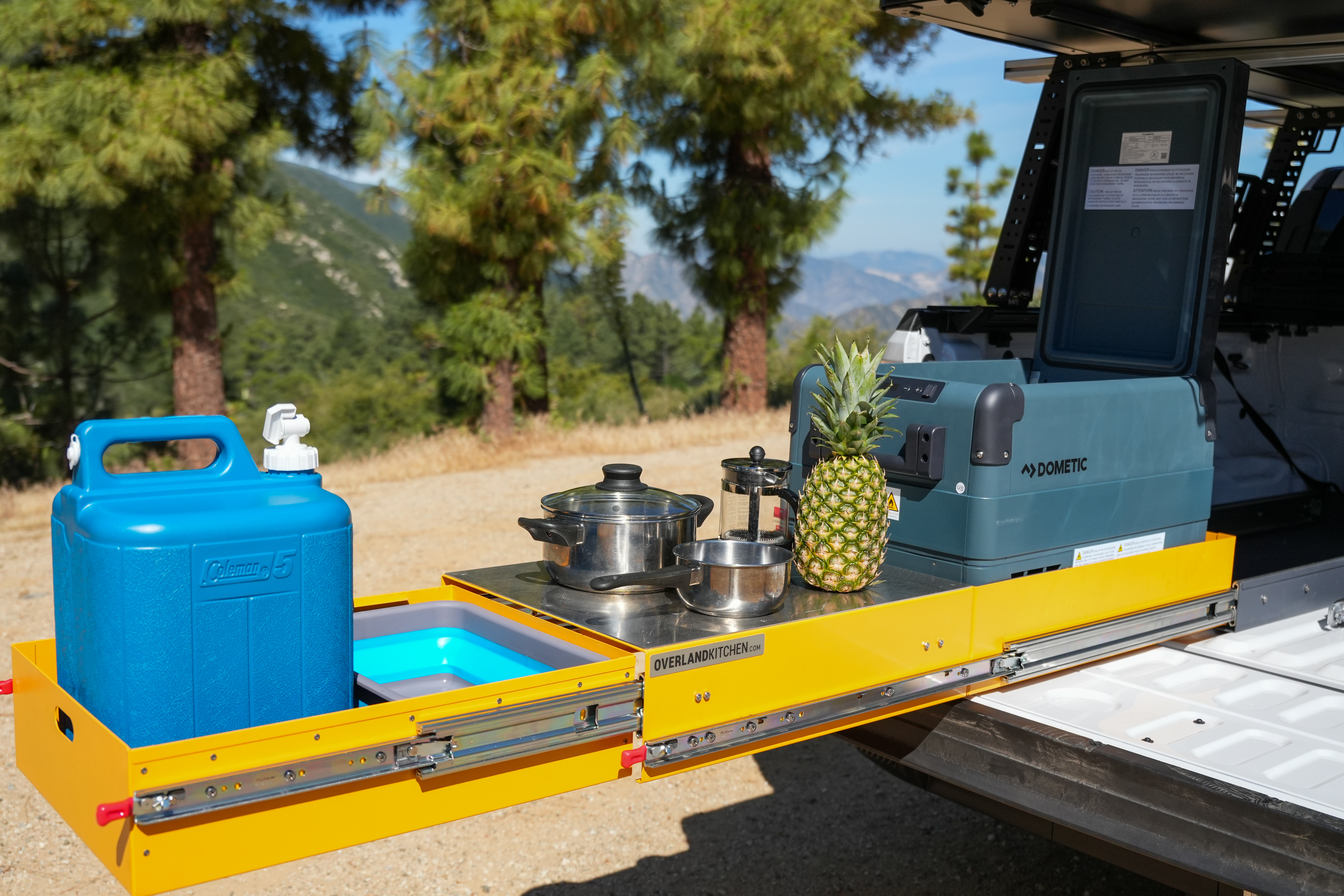 Pull-out camper kitchen setup with a water container, cooking pots, pineapple, and portable fridge in a forested area.