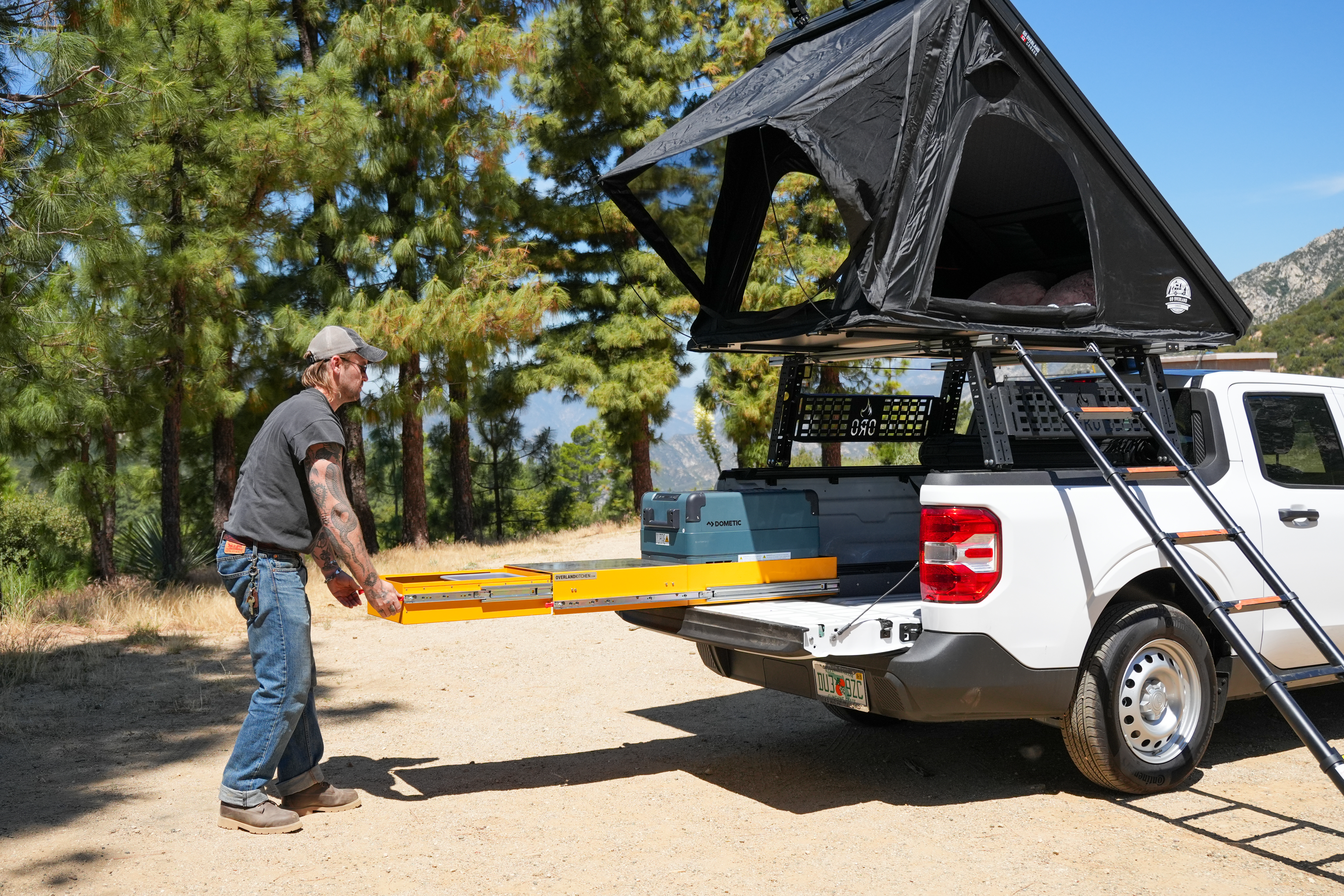 Man pulling out storage drawer from camper van with rooftop tent in a forested outdoor setting.