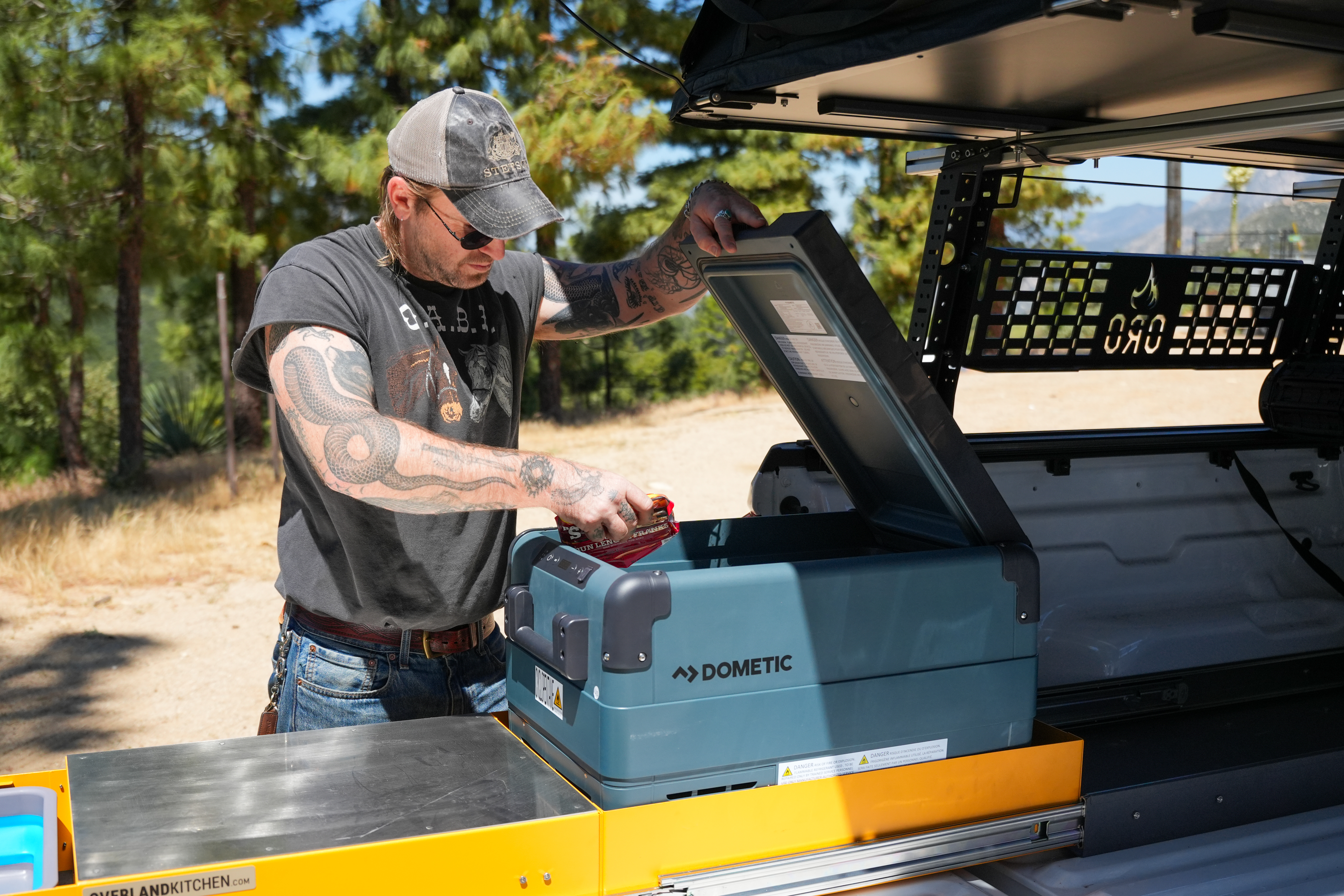 Man opening a Dometic portable cooler in a camper van parked outdoors near trees, under bright daylight.