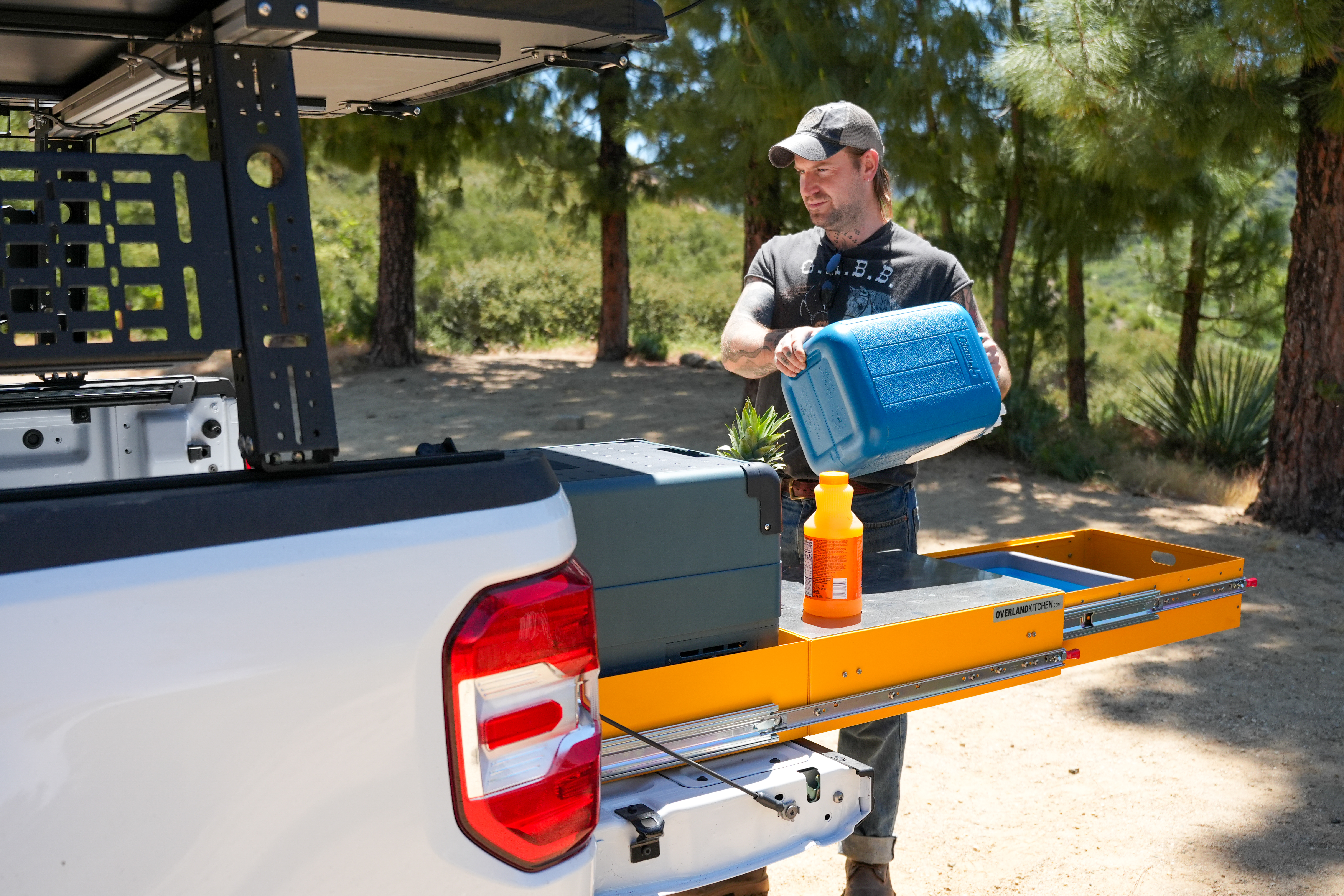 Man pouring water at a truck’s pull-out kitchen setup in a sunny forest campsite.