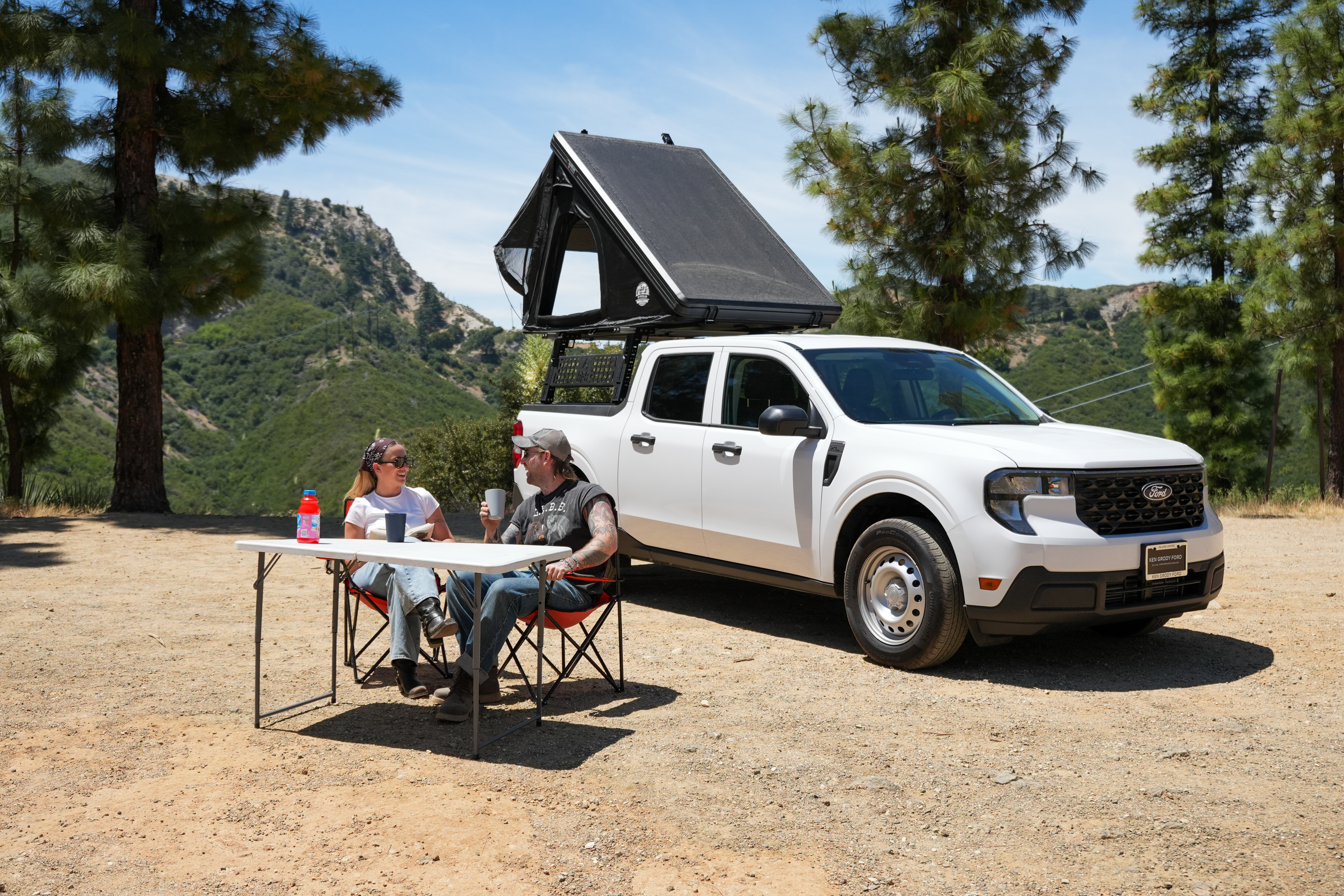 Two people sit at a table beside a campervan with a rooftop tent, surrounded by trees and mountains.
