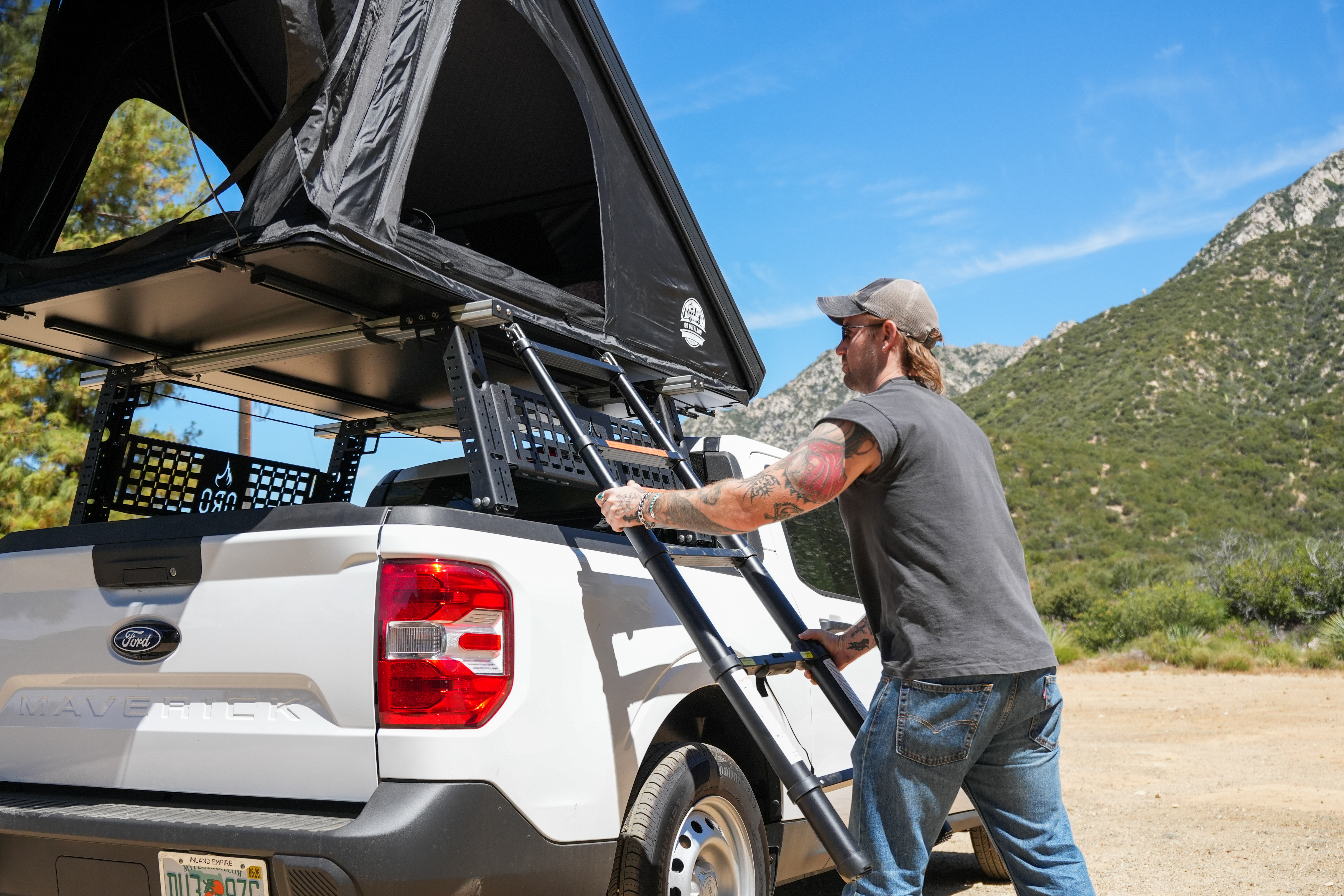 Man setting up rooftop tent on white Ford pickup truck in outdoor mountain setting under blue sky.
