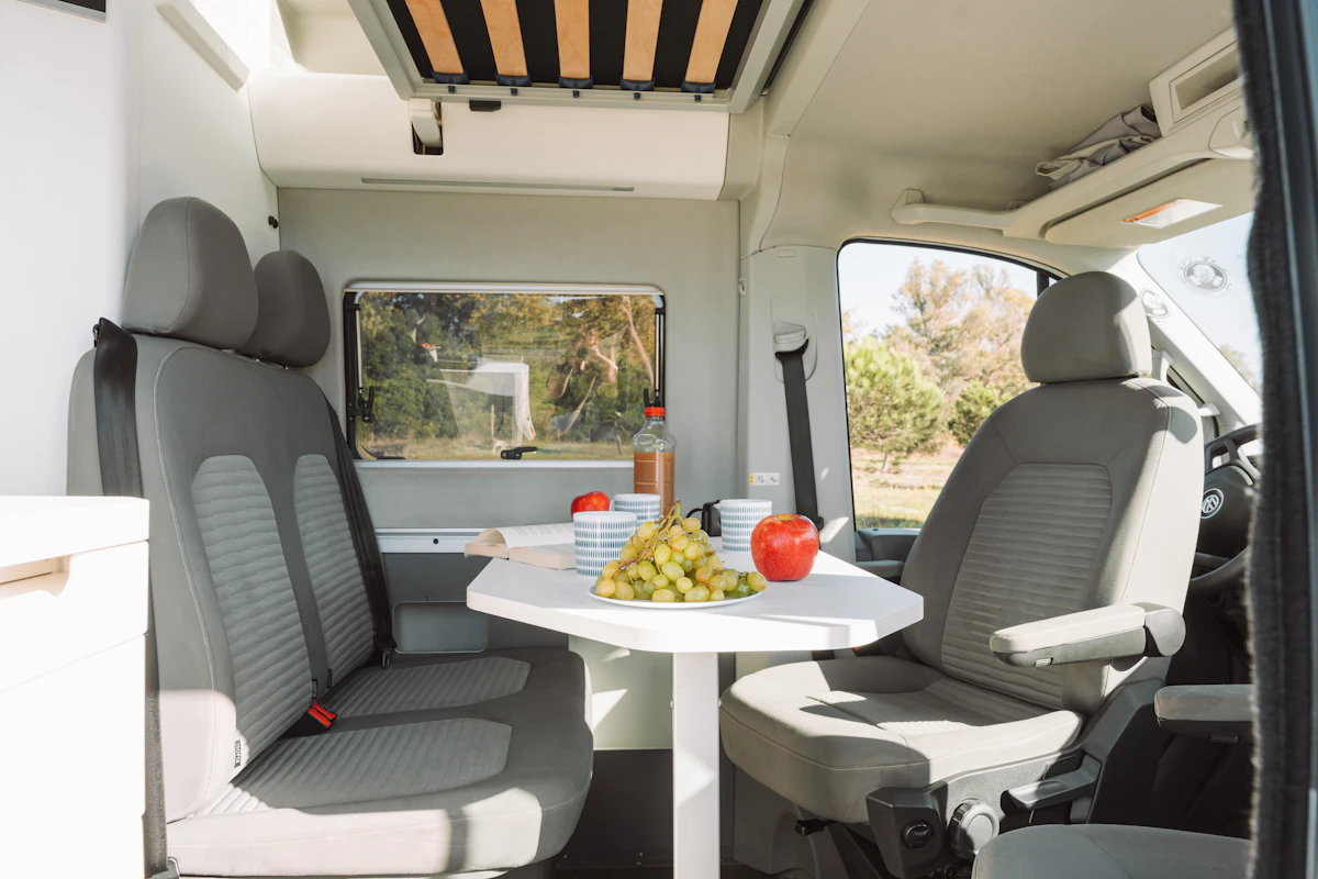 Van interior with two seats, a table, fruit, and a bottle; compact dining area layout.