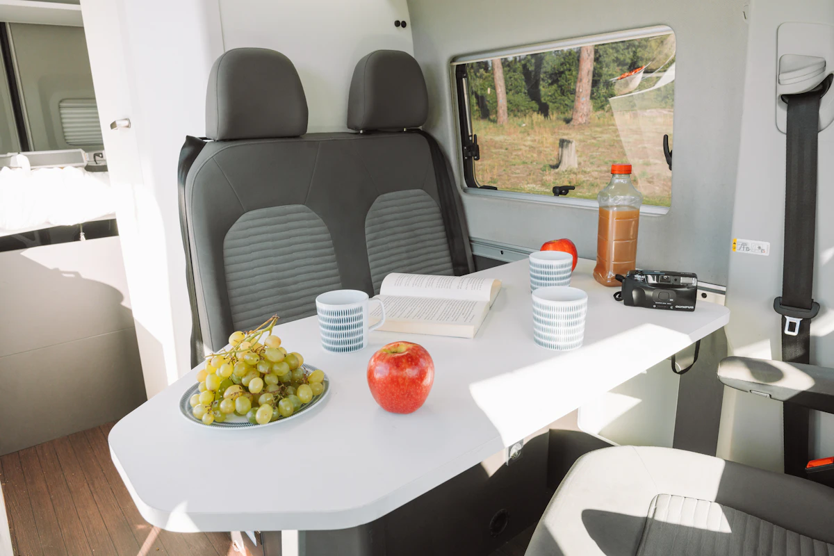 Camper van interior with dining table, two seats, plates, cups, fruit, open book, and side window visible.