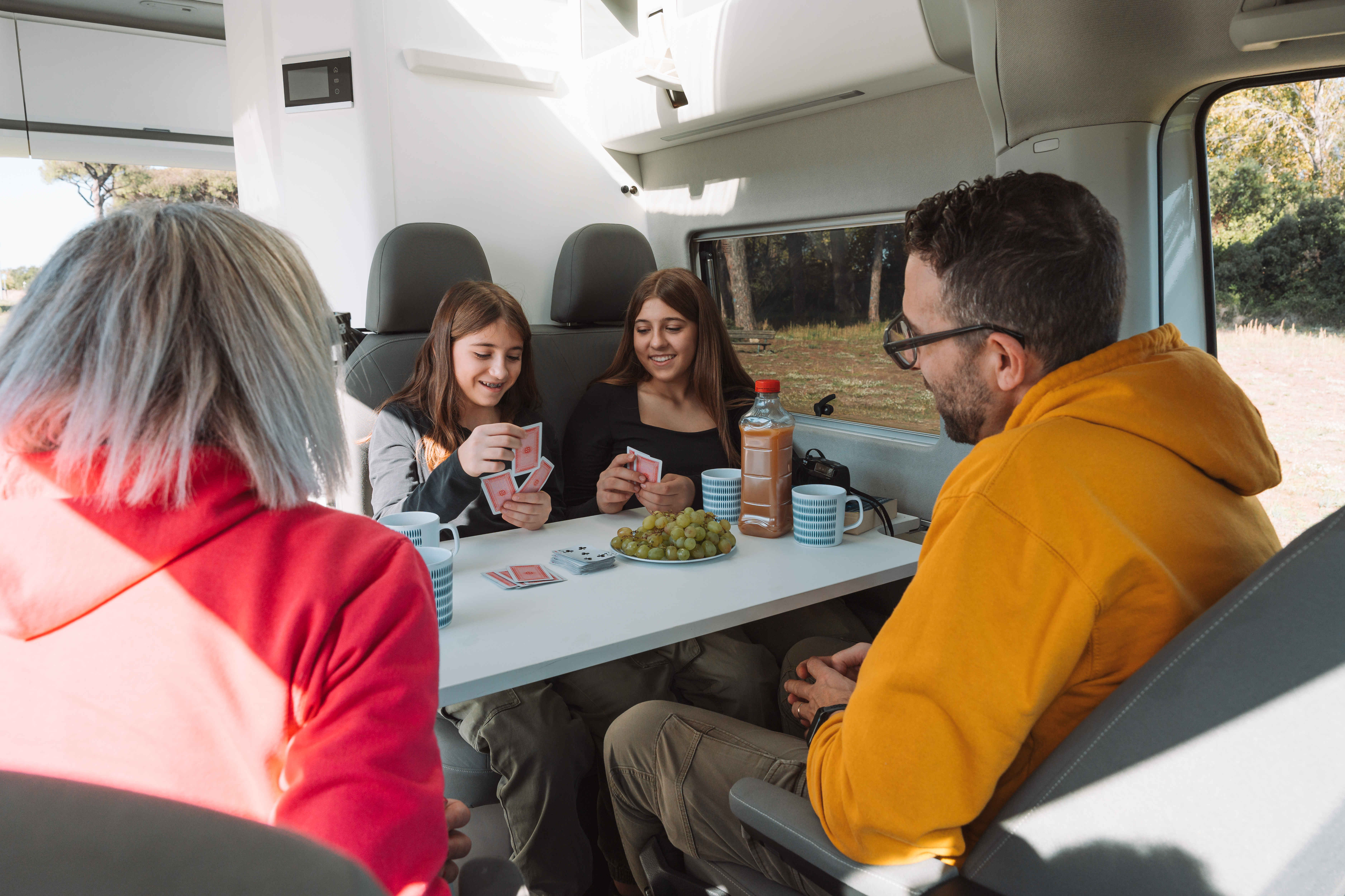 Van interior with dining table, four seats, overhead storage, window, cups, and playing cards on the table.