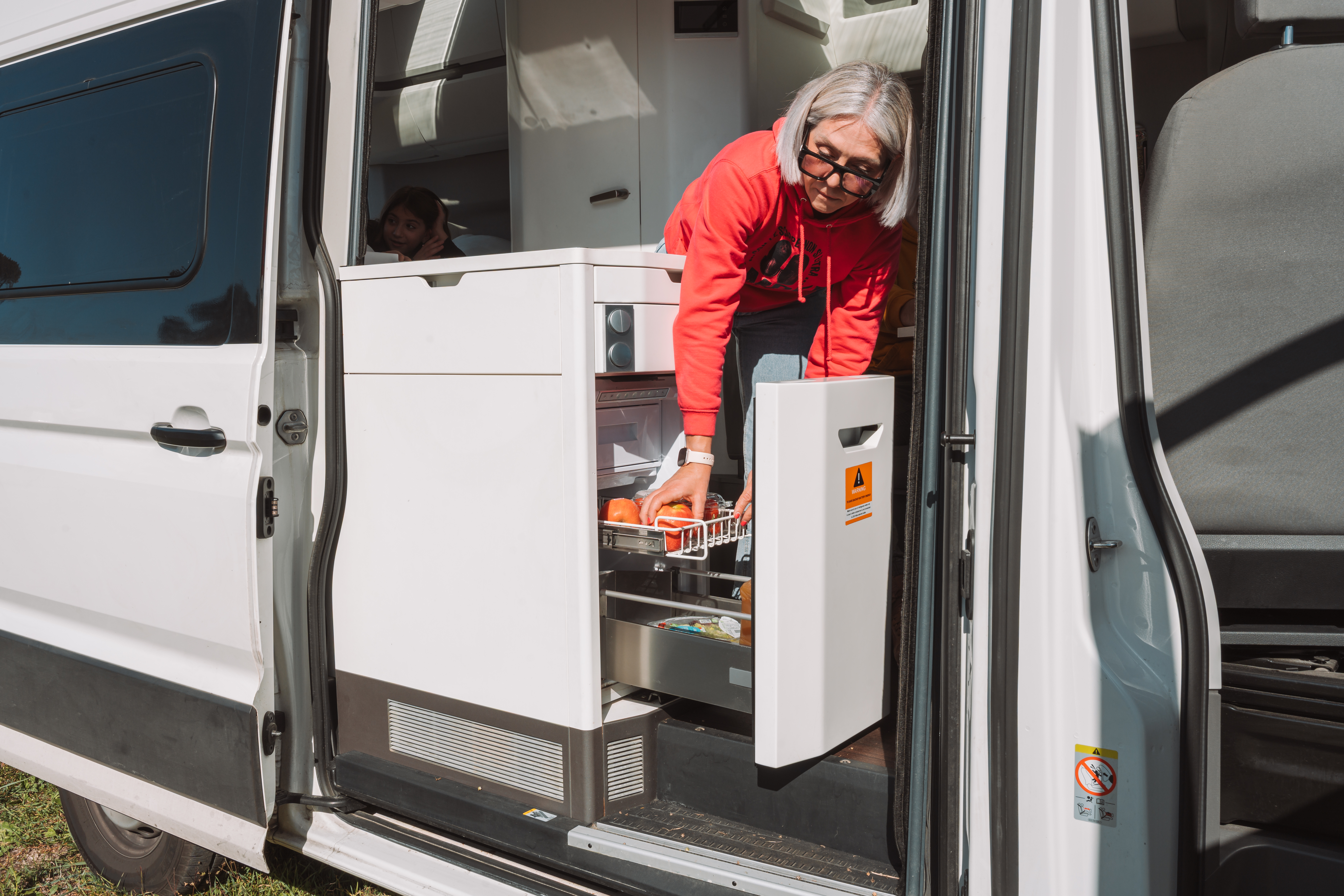 Van interior with compact kitchen area, pull-out fridge drawer, and white cabinetry visible by the open side door.