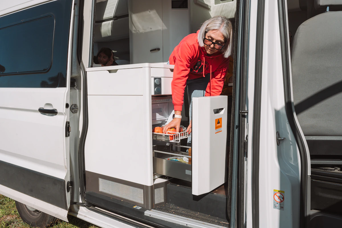 Van interior with compact kitchen area, pull-out fridge drawer, and white cabinetry visible by the open side door.