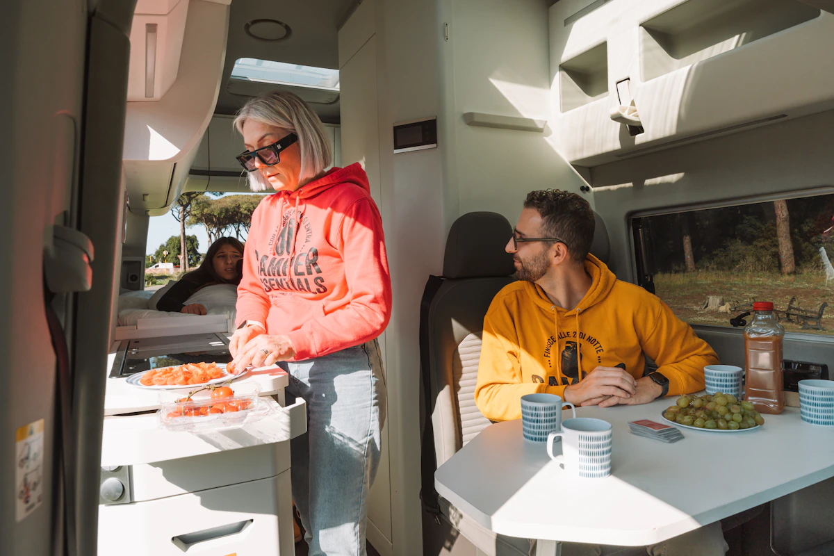 Camper van interior with kitchenette, dining table, seating, cabinets, and kitchen equipment visible.