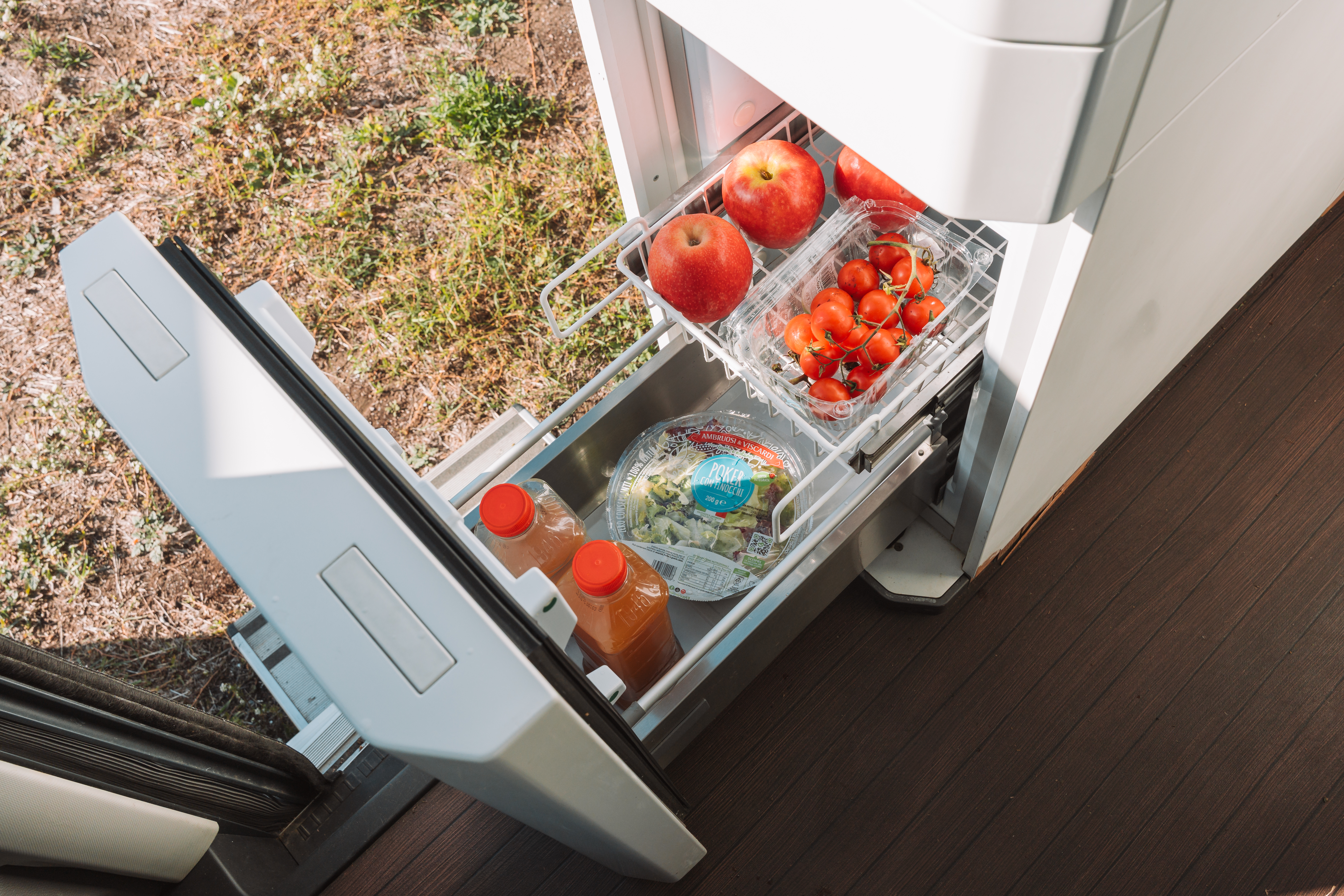 Van interior with open drawer fridge containing apples, tomatoes, salad, and juice bottles next to wooden flooring.