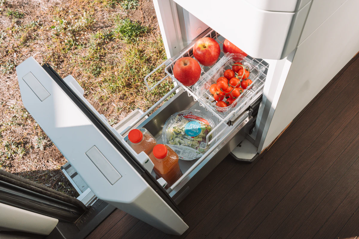 Van interior with open drawer fridge containing apples, tomatoes, salad, and juice bottles next to wooden flooring.