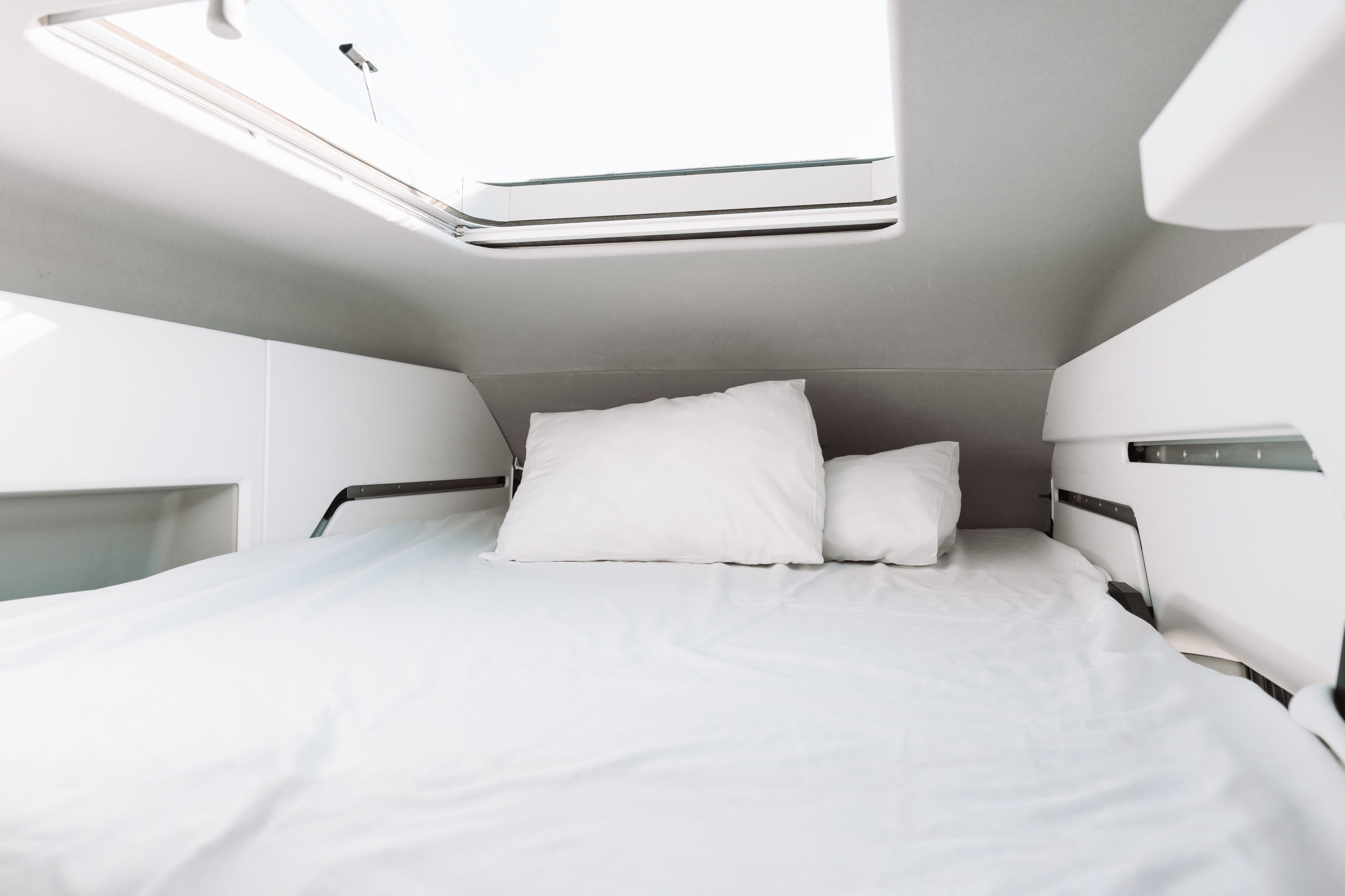 Double bed with white linen and pillows beneath skylight, surrounded by white walls and storage shelves inside van.
