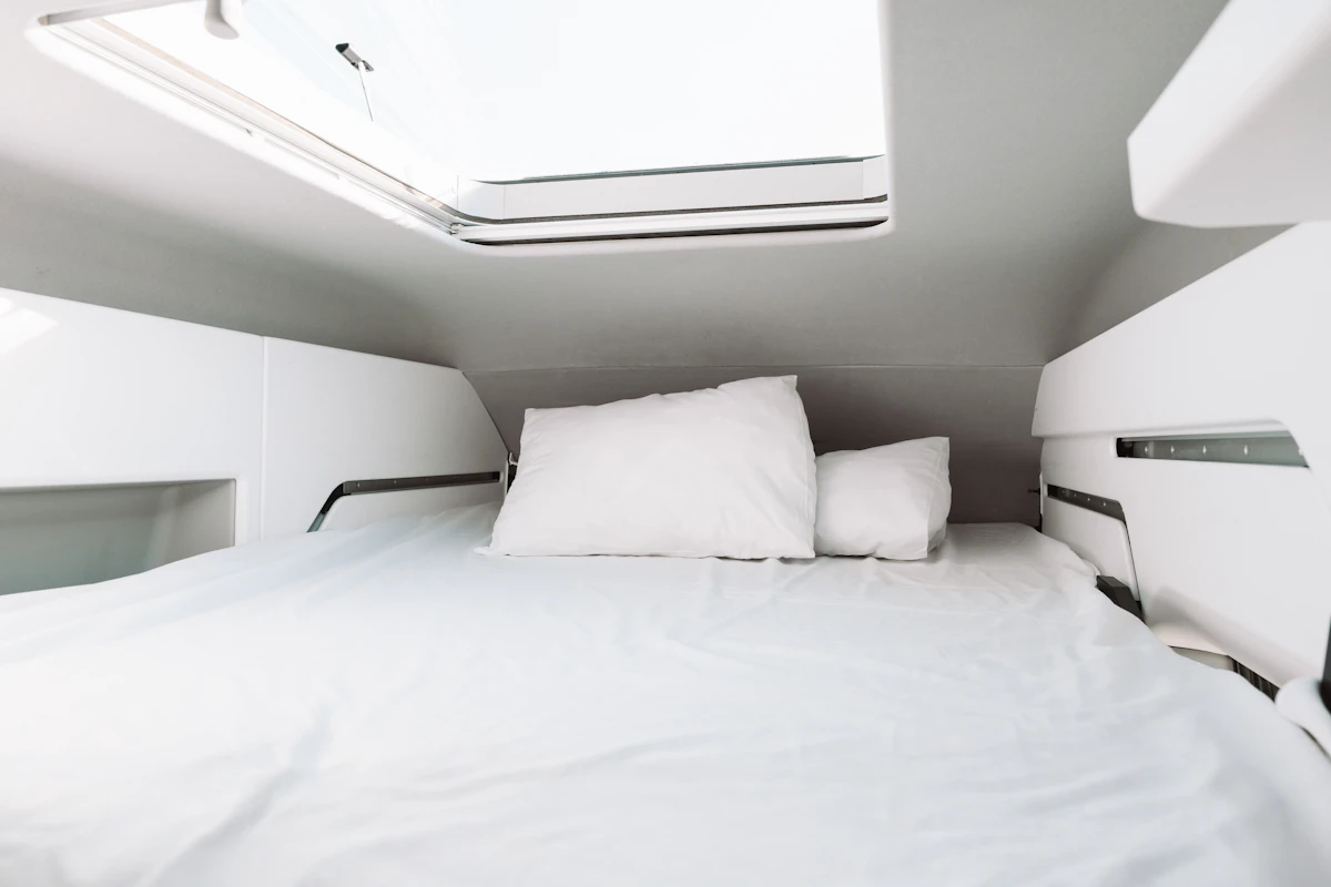 Double bed with white linen and pillows beneath skylight, surrounded by white walls and storage shelves inside van.