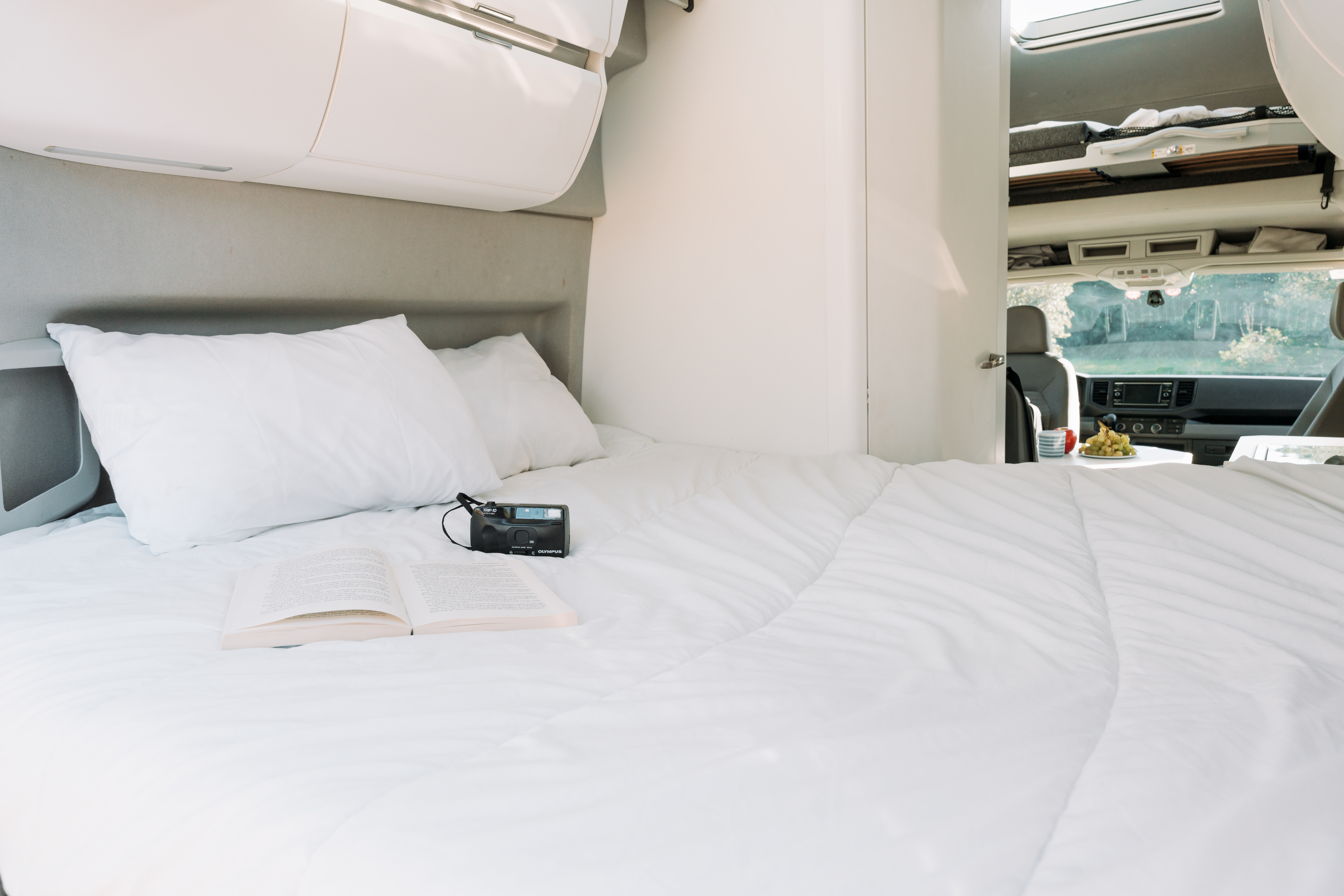 Van interior with large bed, pillows, overhead storage, front cabin, and upper sleeping area visible.