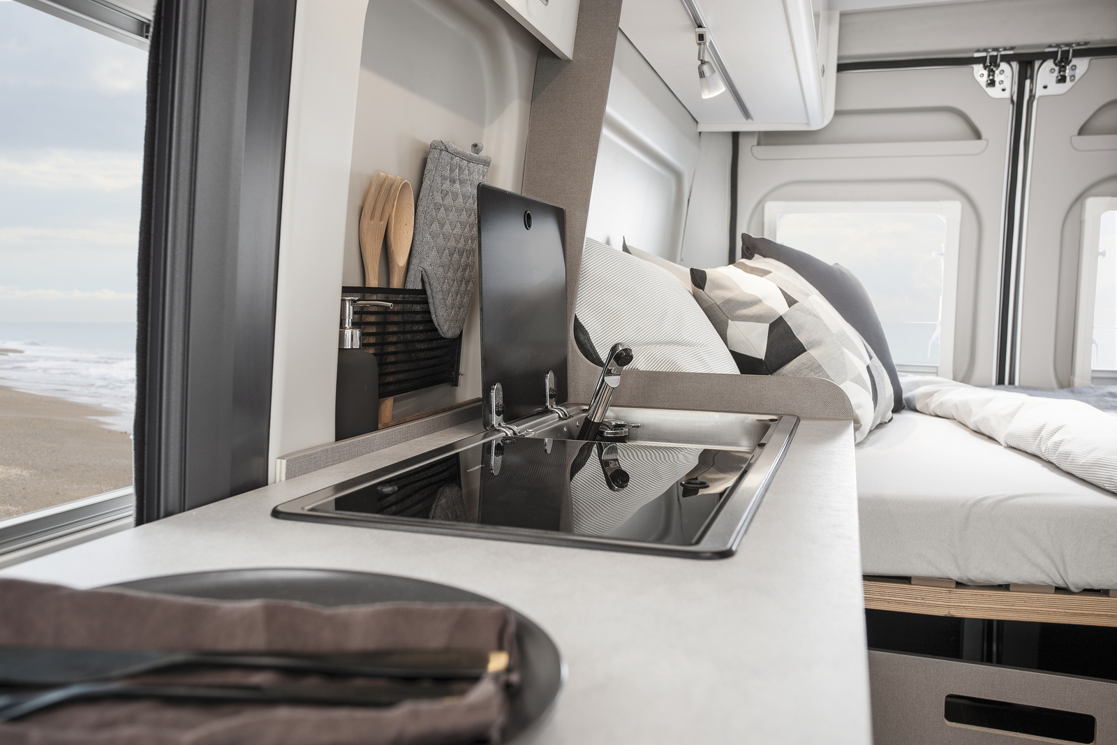 Van interior with kitchen sink, stove, cooking utensils, and a bed in the background.