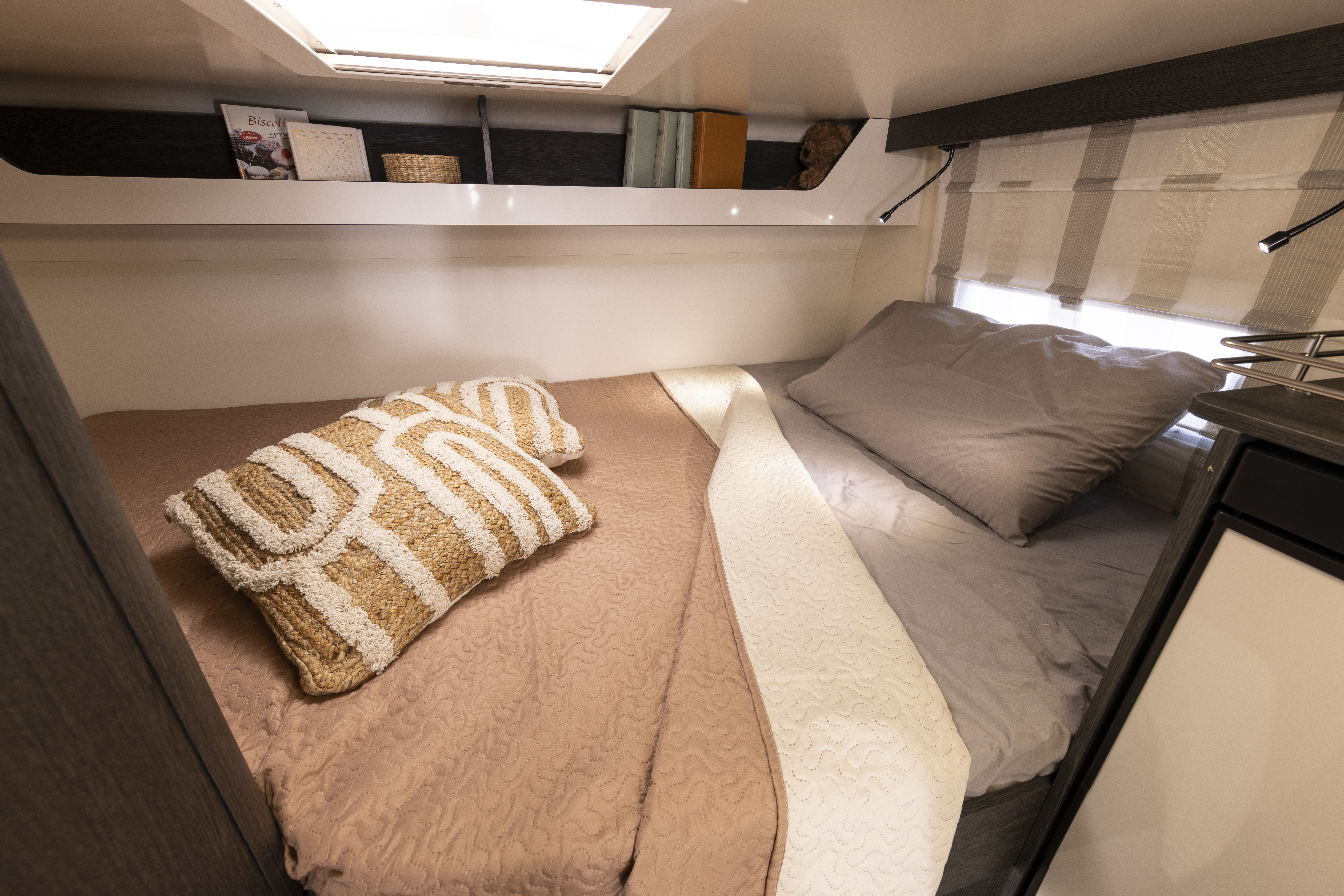 Double bed with bedding, pillows, side shelf, window blinds, and overhead storage inside a campervan.