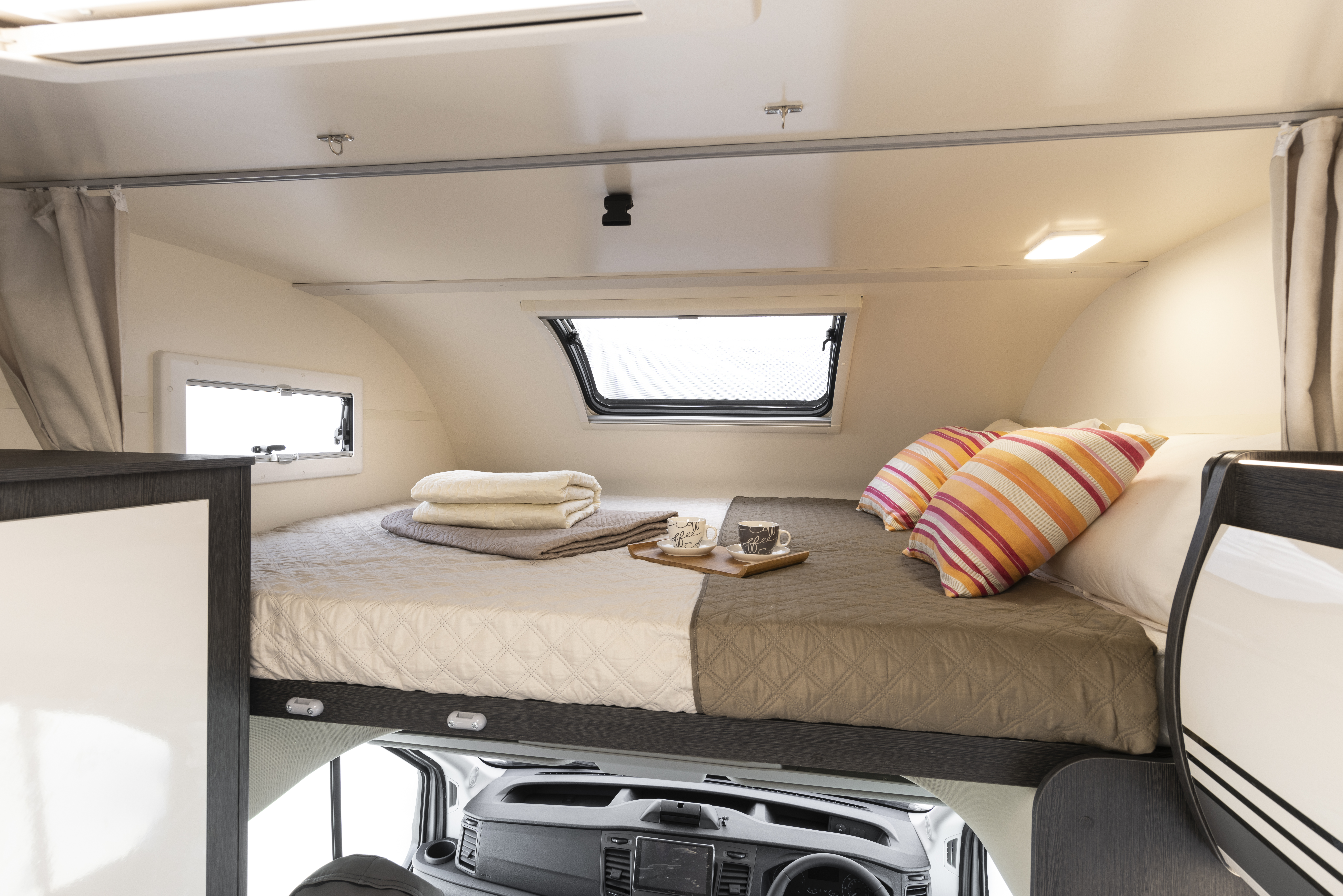 Overhead bed area with pillows, folded towels, tray with cups, and windows above campervan driver’s cabin.