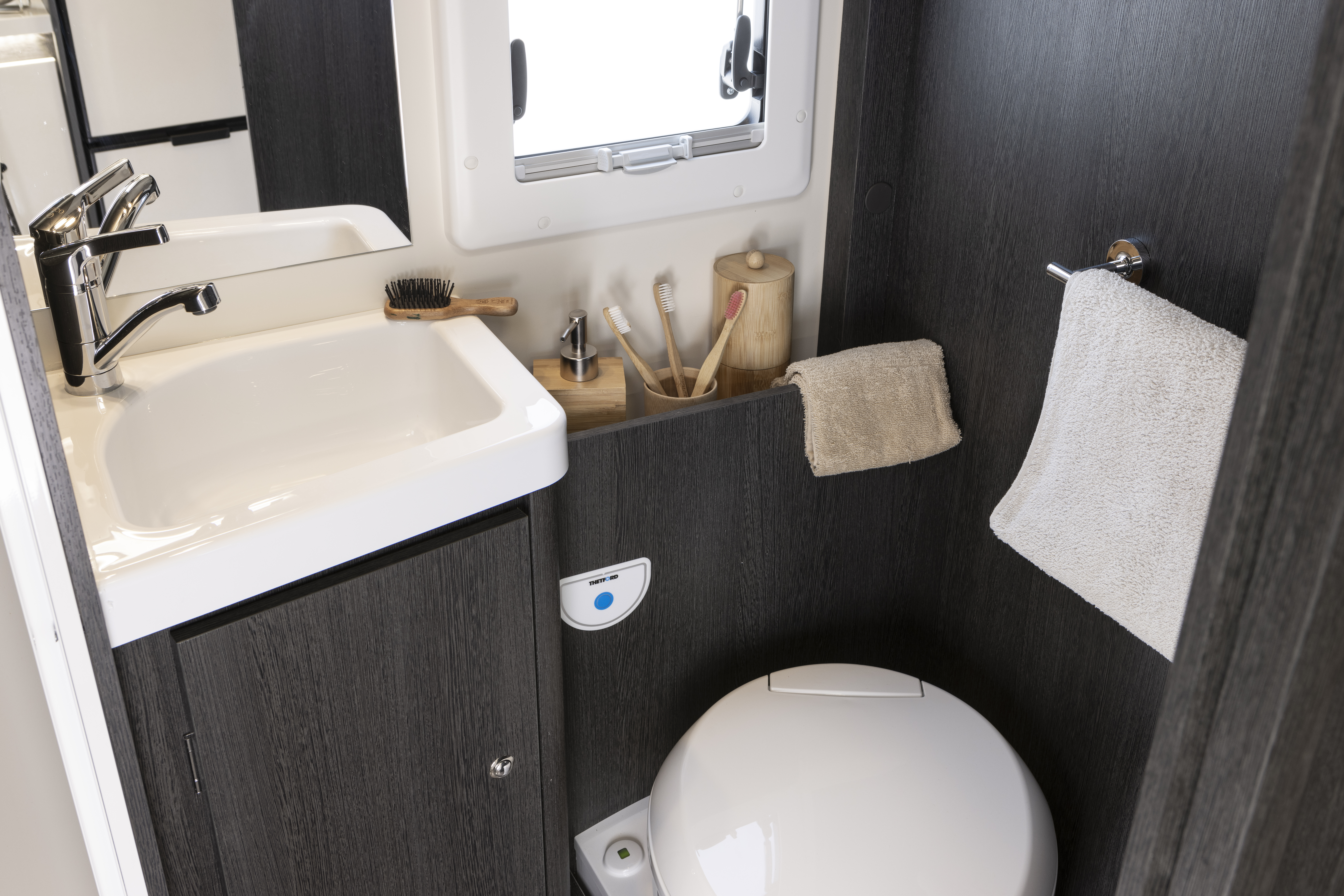Compact van bathroom with sink, toilet, towel rail, toiletries, and small window on dark wood cabinetry.