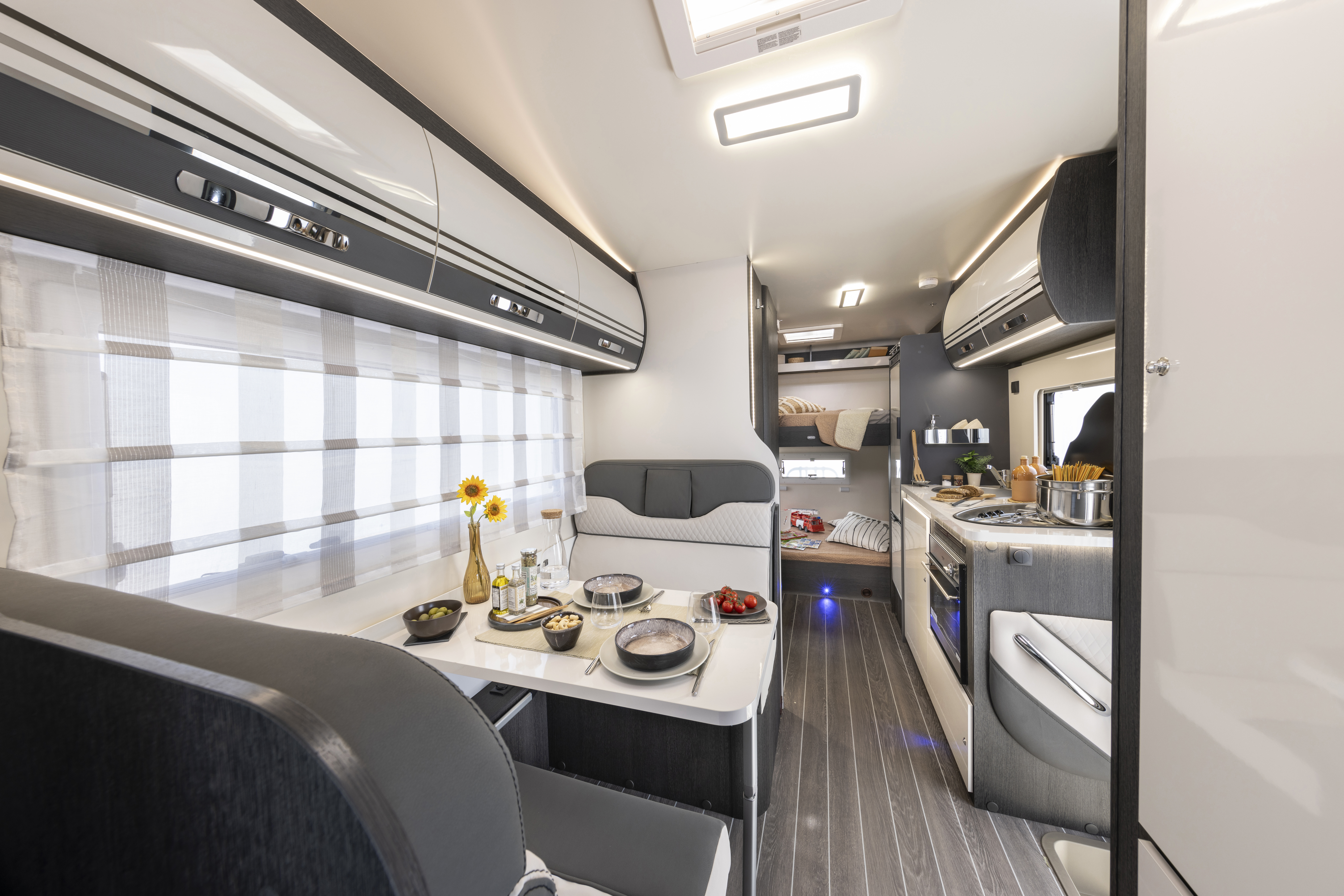 Dining area, kitchen, and sleeping space inside campervan with table, seating, cabinets, stove, and bed visible.