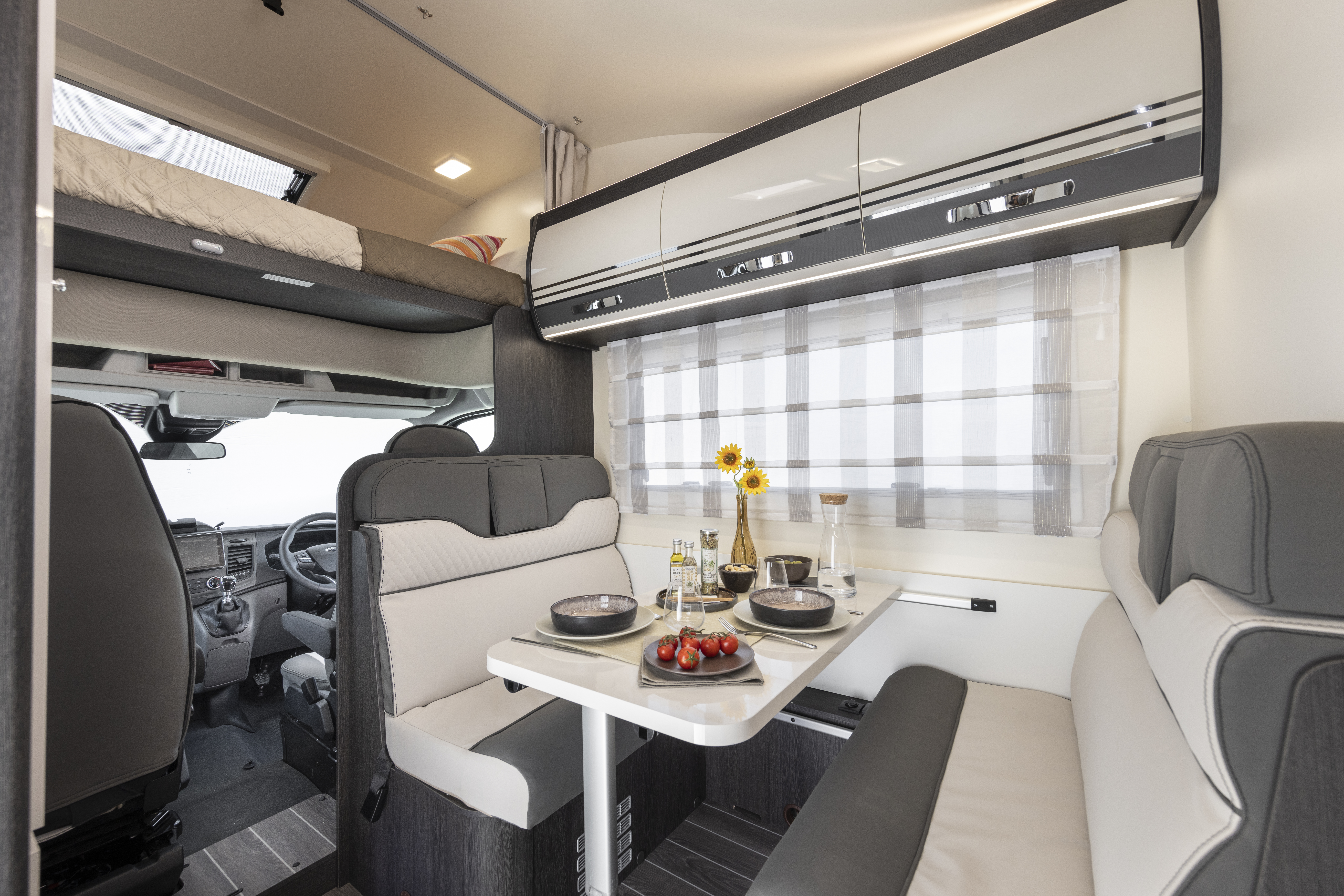 Van interior with dinette, two bench seats, fold-out table, overhead bed, storage cabinets, and window with curtain.