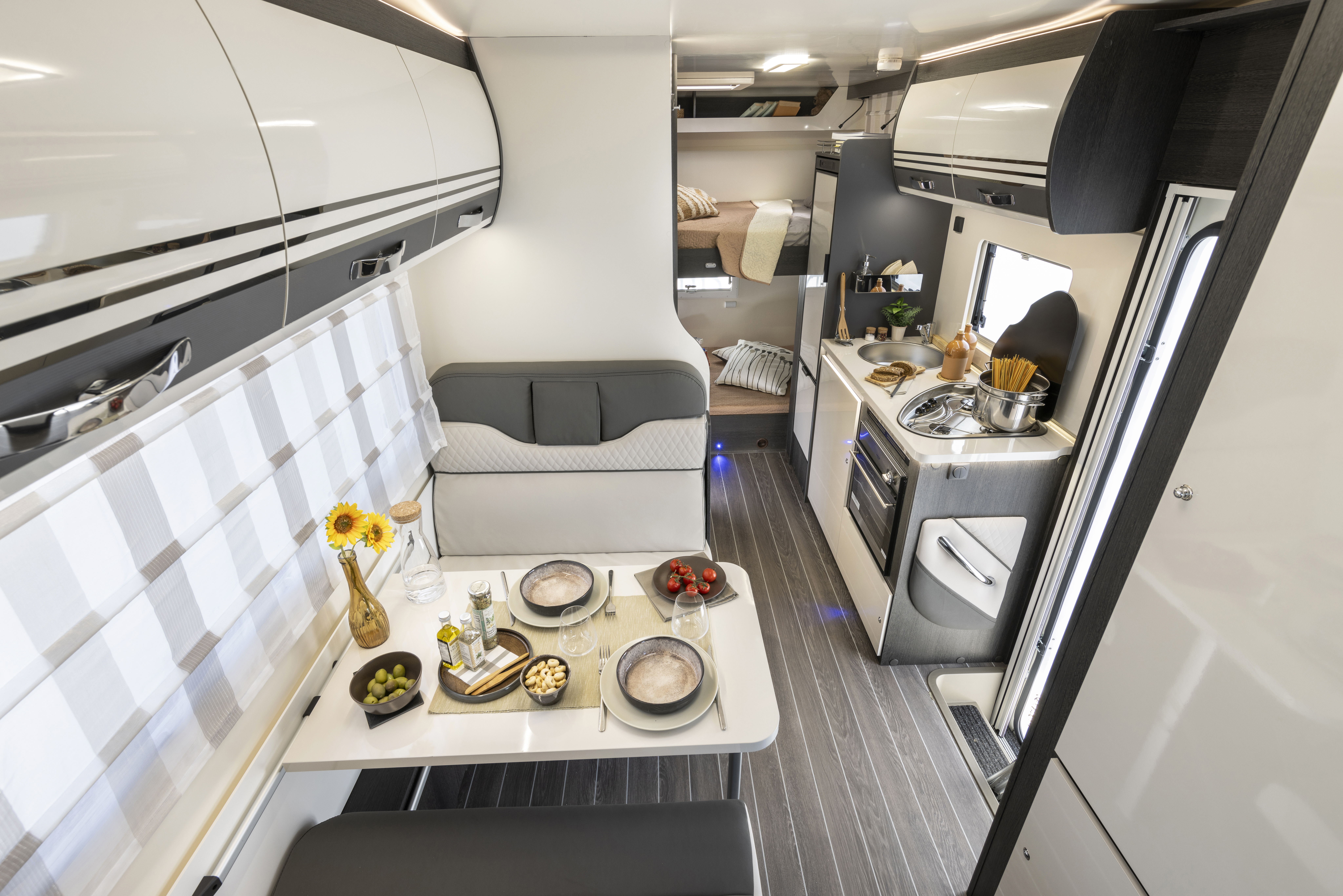 Modern campervan interior with dining table, bench seating, kitchenette, overhead storage, and visible sleeping area in the back.
