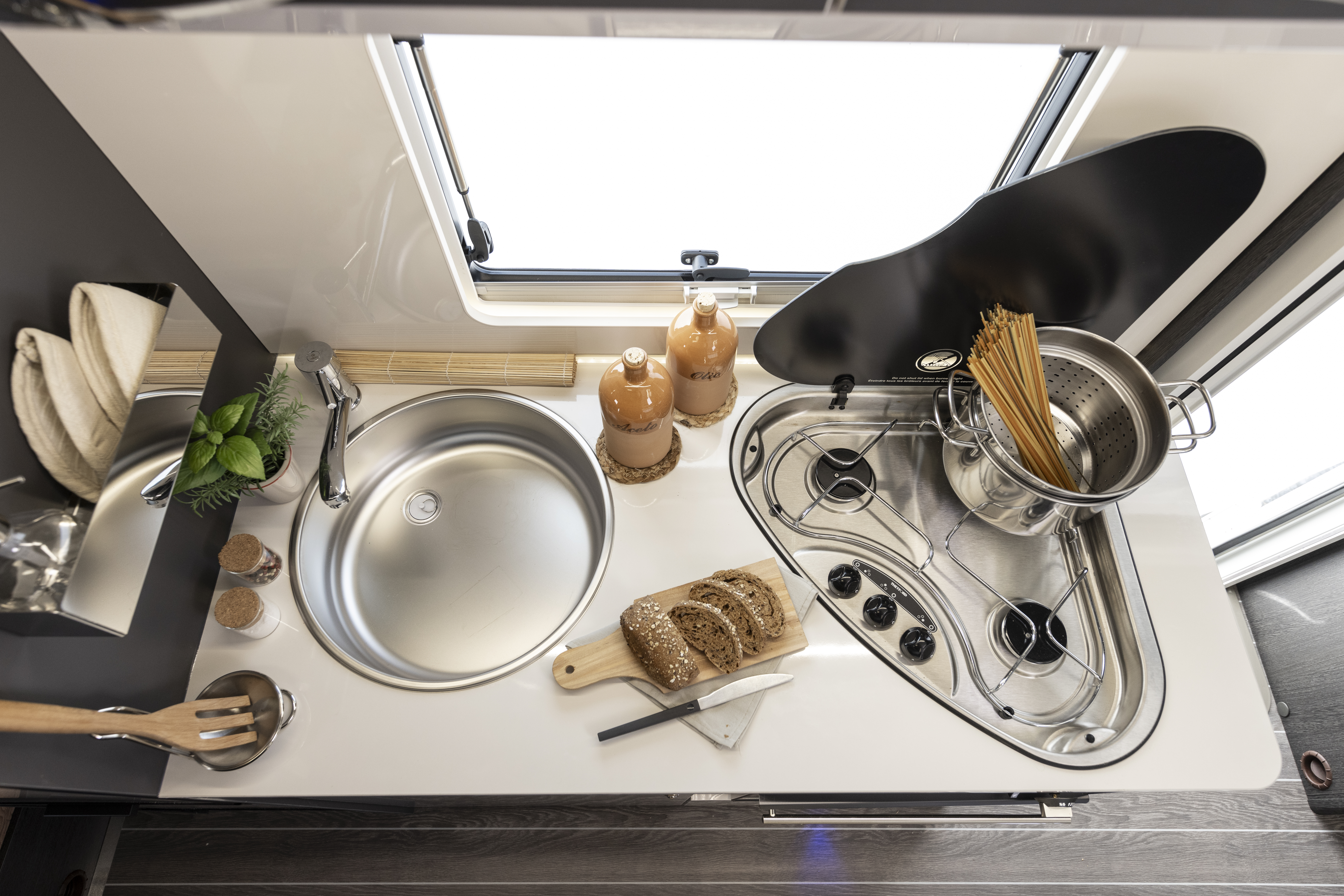 Compact van kitchen with sink, two-burner stove, pot, utensils, bread, and window above the countertop.
