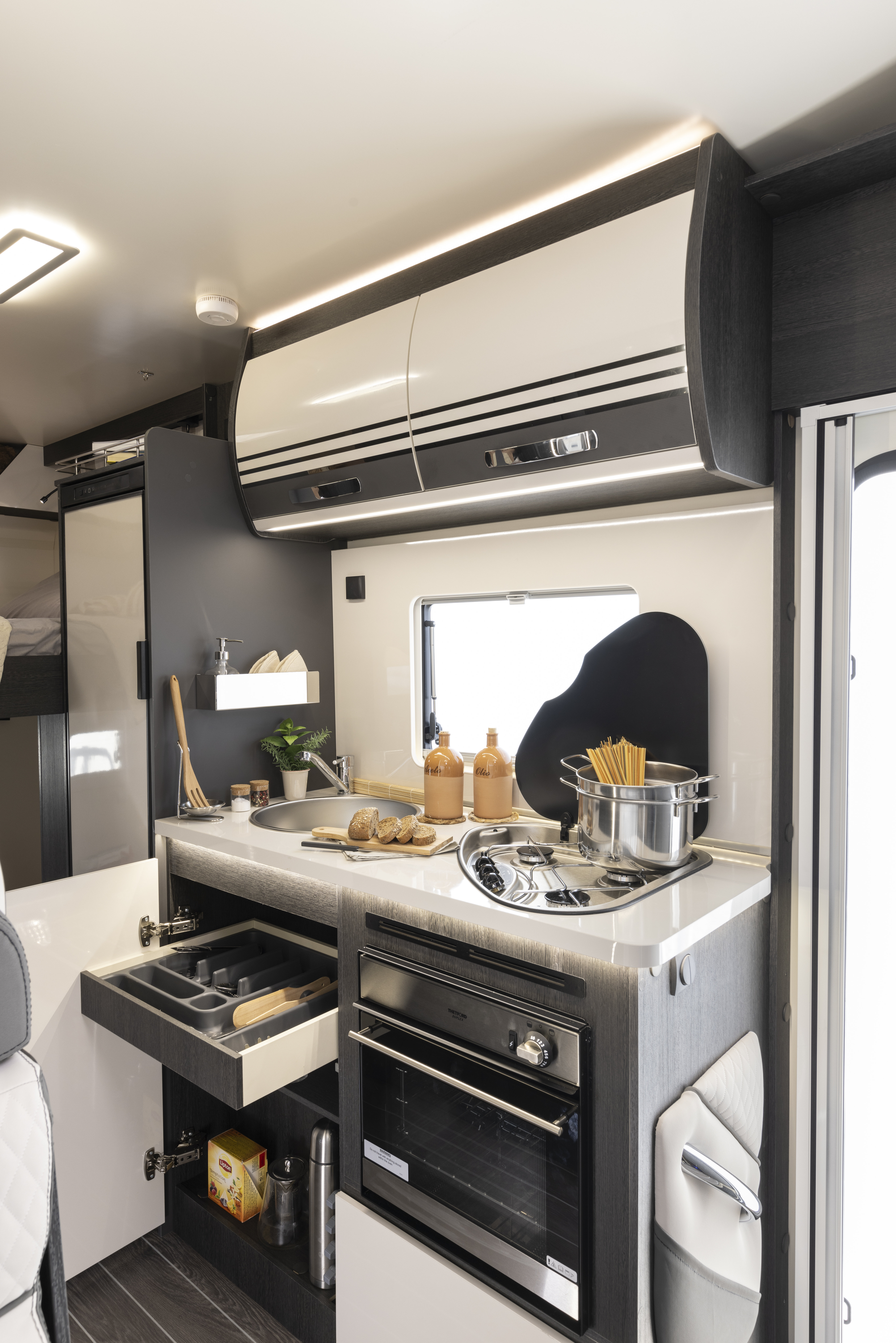 Compact campervan kitchen interior with stove, sink, oven, storage drawers, and kitchen utensils visible.