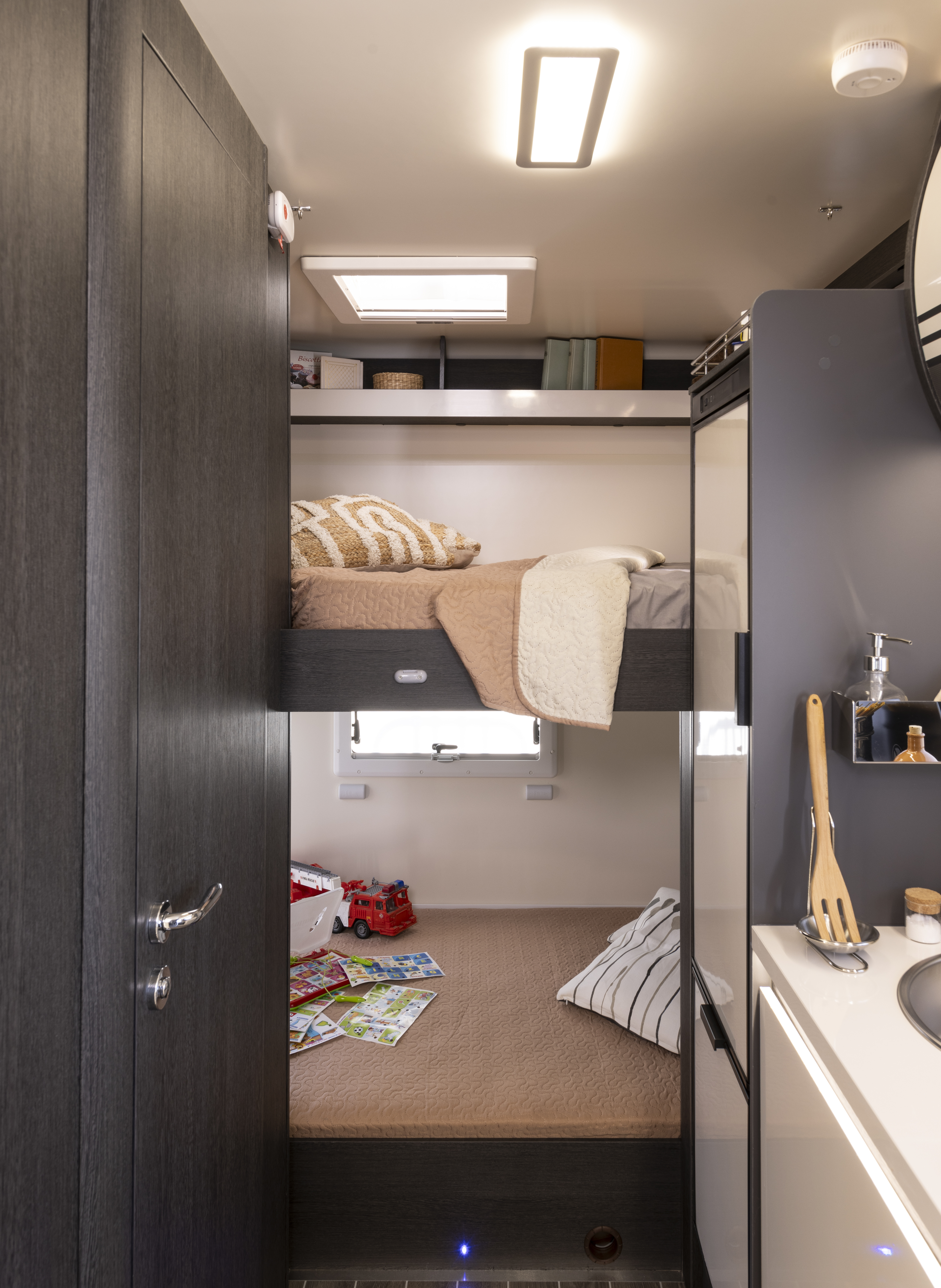 Van interior with bunk beds, window, books, toy truck, striped cushion, and part of kitchenette visible.