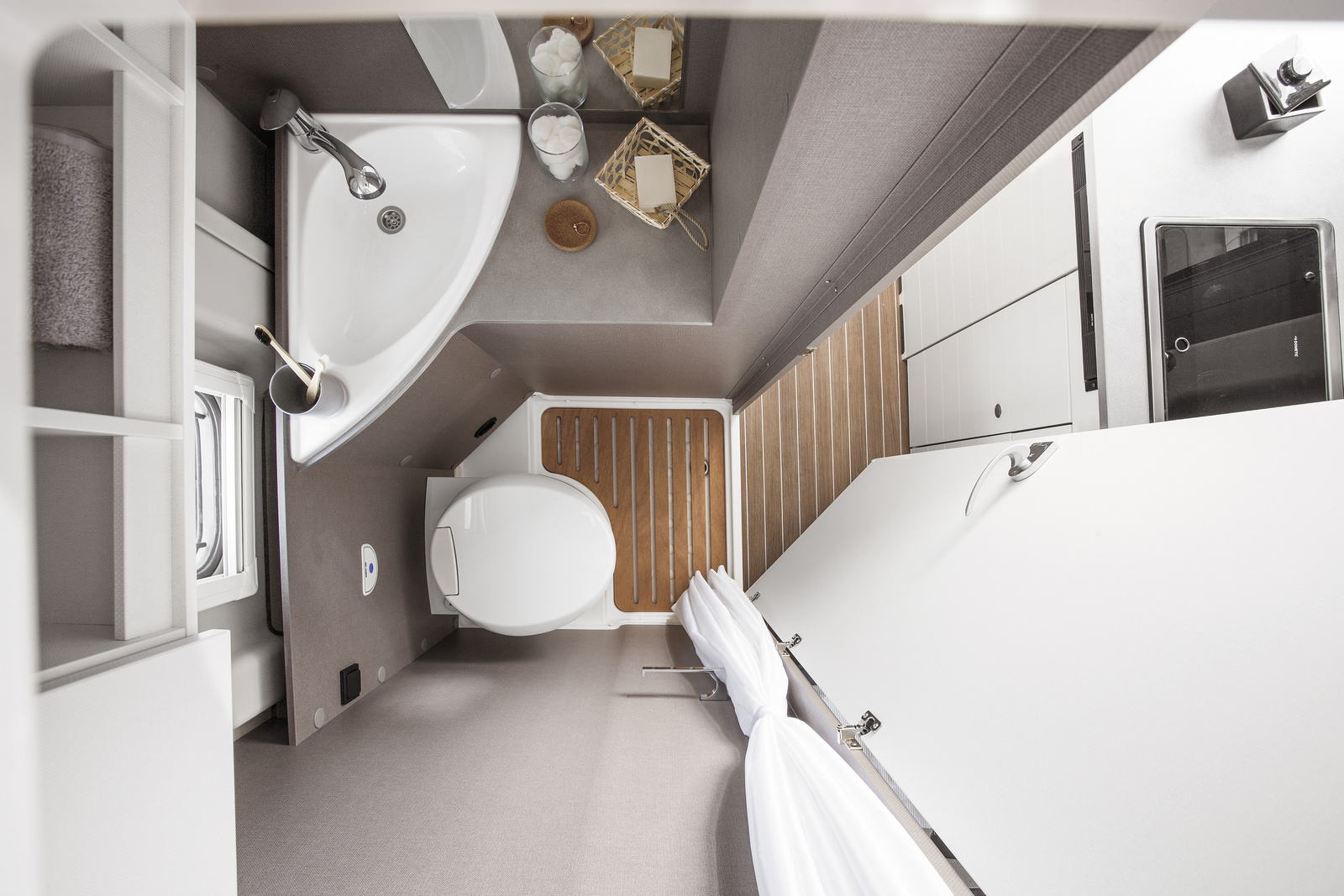 Compact van bathroom with toilet, corner sink, shelves, shower curtain, wooden floor mat, and storage compartments.