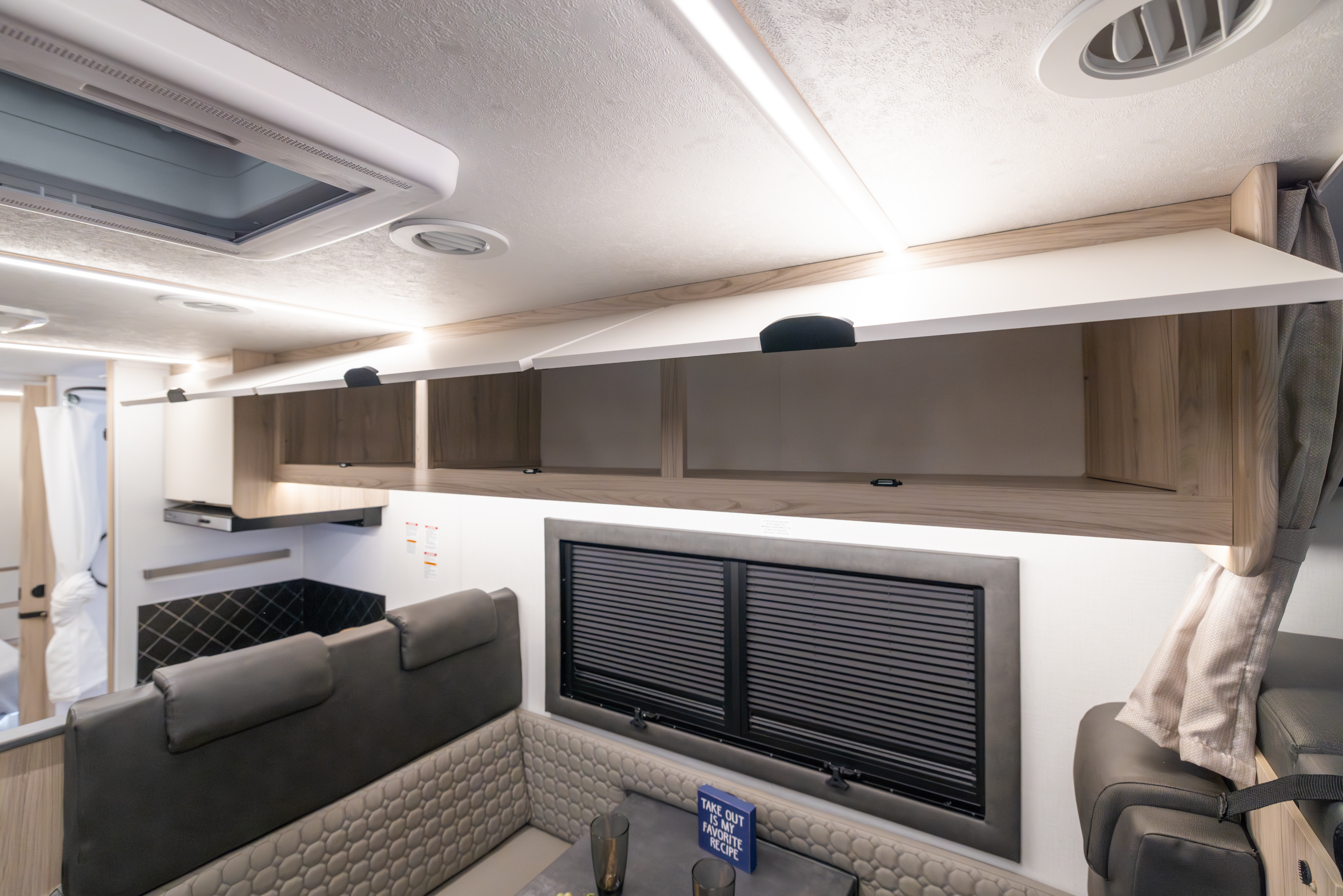 Van interior with open overhead cabinets, dining area seating, table, kitchenette, and two closed windows.