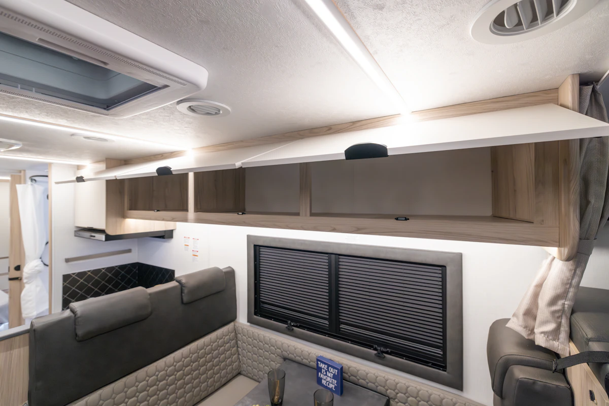 Van interior with open overhead cabinets, dining area seating, table, kitchenette, and two closed windows.
