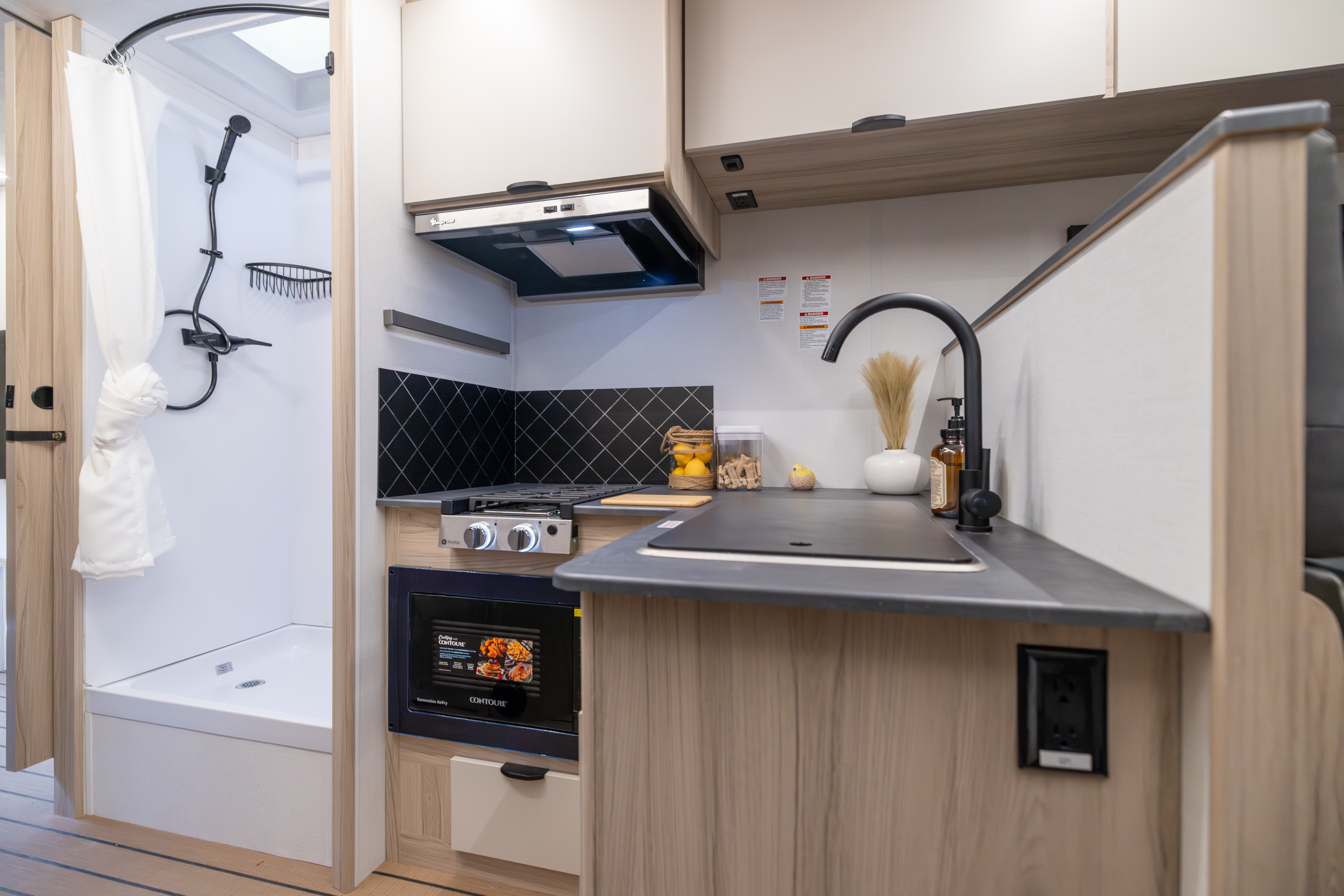 Compact campervan kitchen with stove, microwave, sink, cupboards, and adjacent shower visible on the left side.