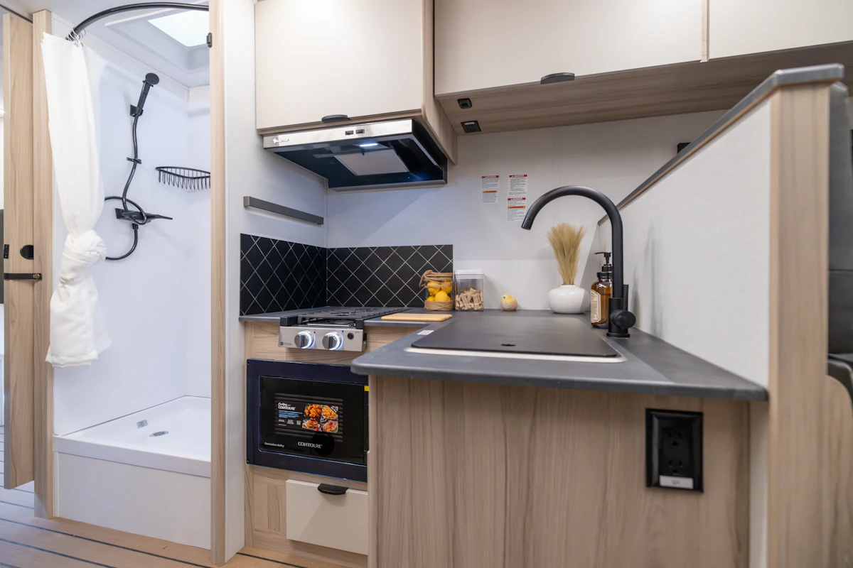 Compact campervan kitchen with stove, microwave, sink, cupboards, and adjacent shower visible on the left side.