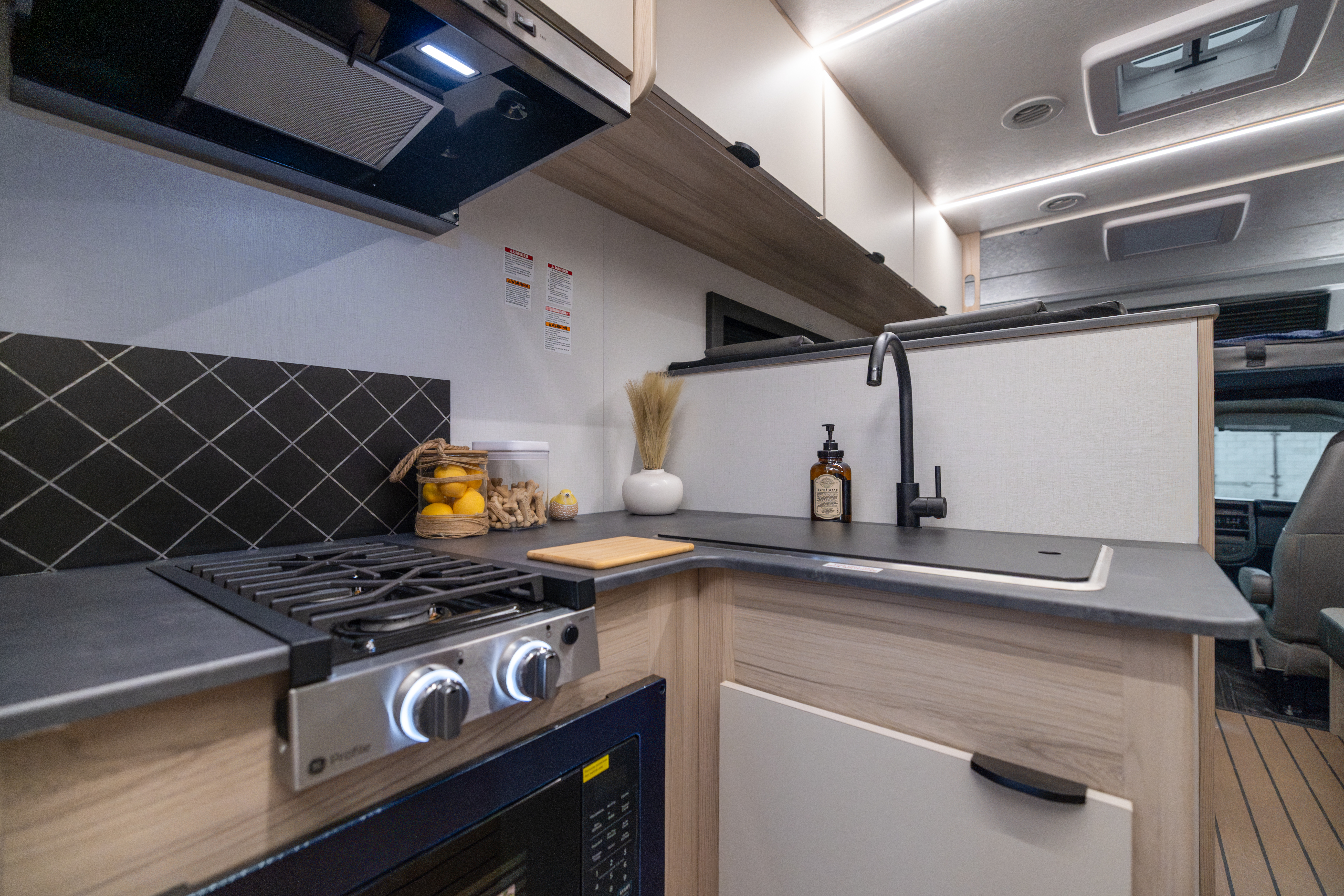 Compact van kitchen with stove, sink, countertop, oven, storage cabinets, and various kitchen accessories and equipment.