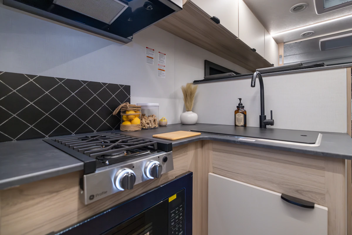 Compact van kitchen with stove, sink, countertop, oven, storage cabinets, and various kitchen accessories and equipment.