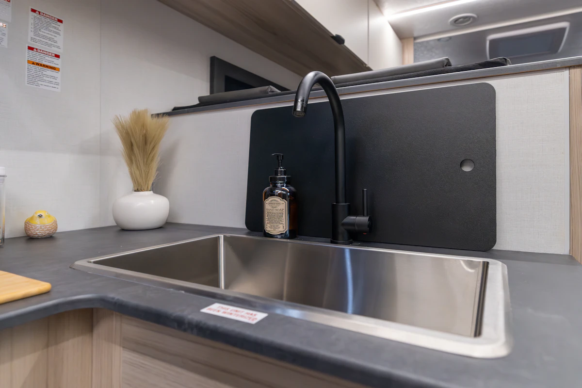 Van kitchen interior with large sink, countertop, faucet, soap dispenser, storage shelves, and small decorative items visible.