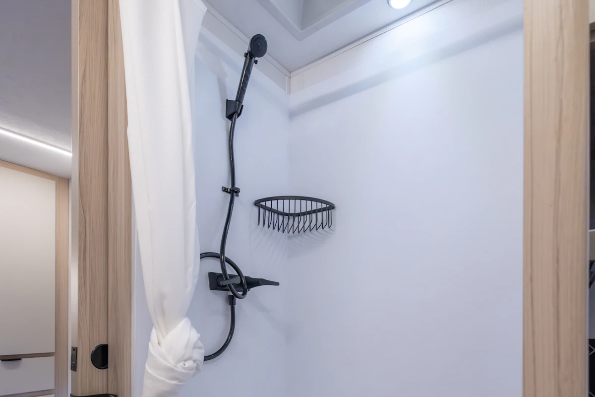Van interior showing compact shower with curtain, black showerhead, soap basket, and overhead lighting.
