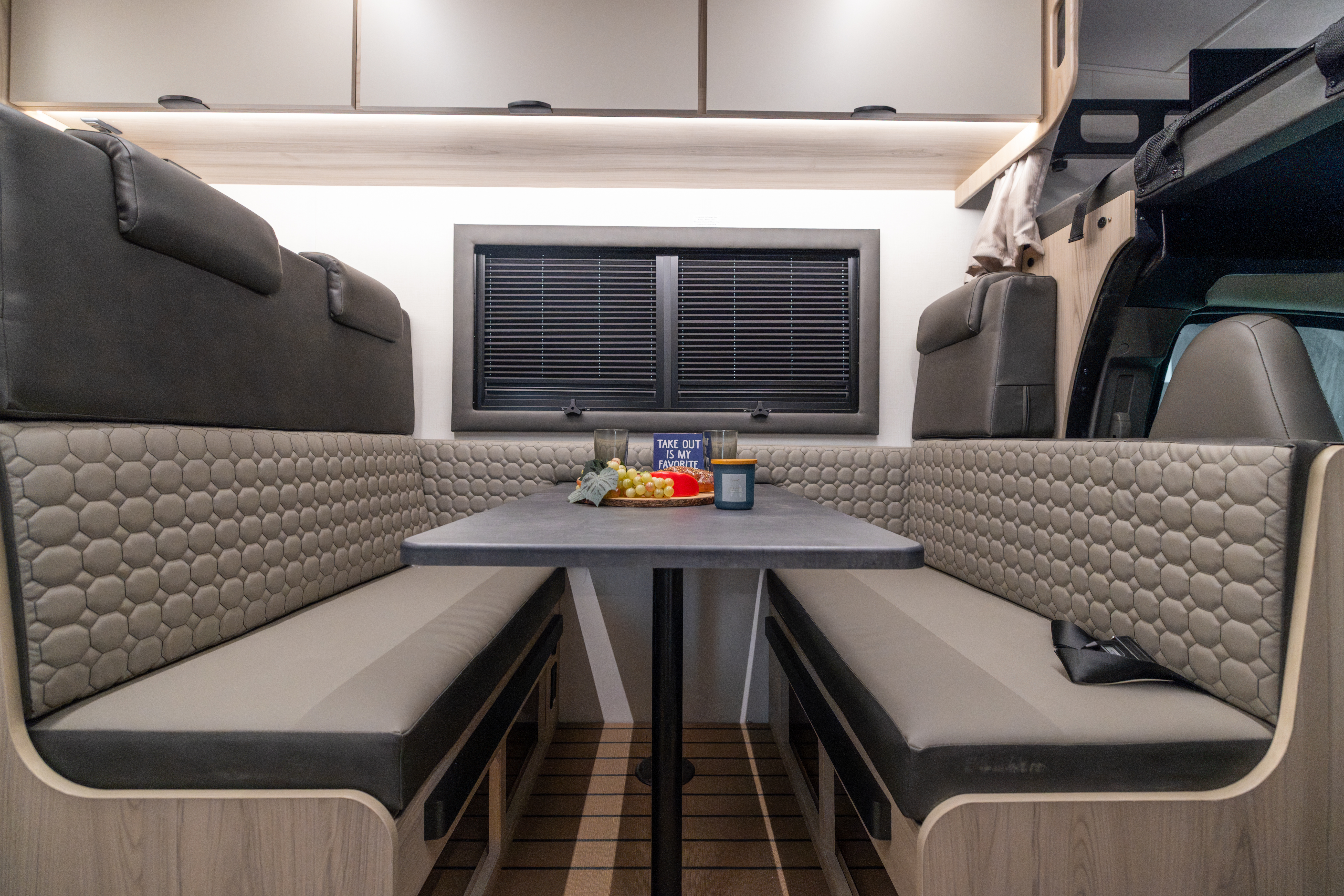 Van interior with two parallel benches, central table, window with blinds, overhead cabinets, and cushioned seating.