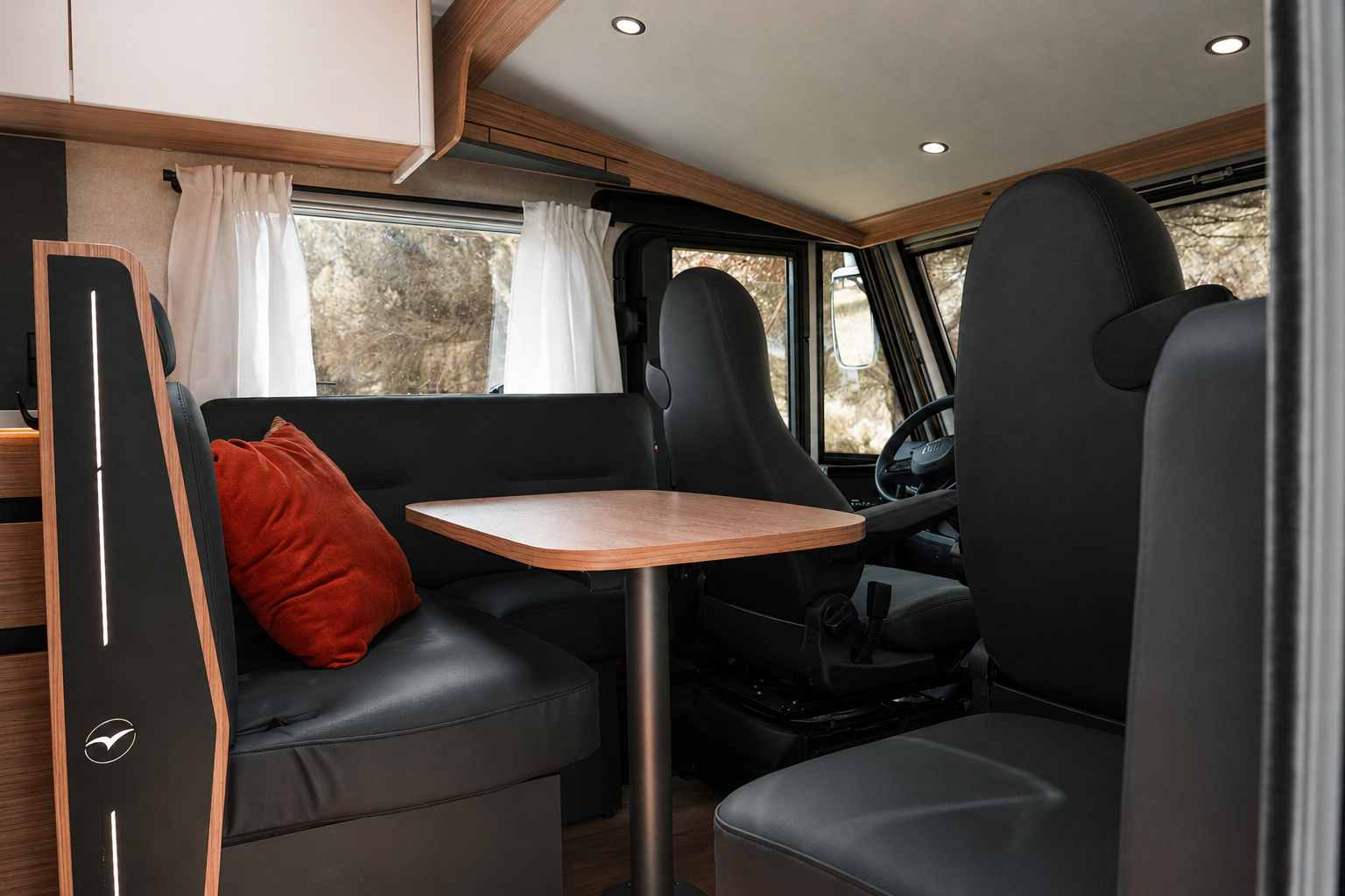 Van interior with front seats, dining table, side bench, overhead cabinets, and window curtains visible.