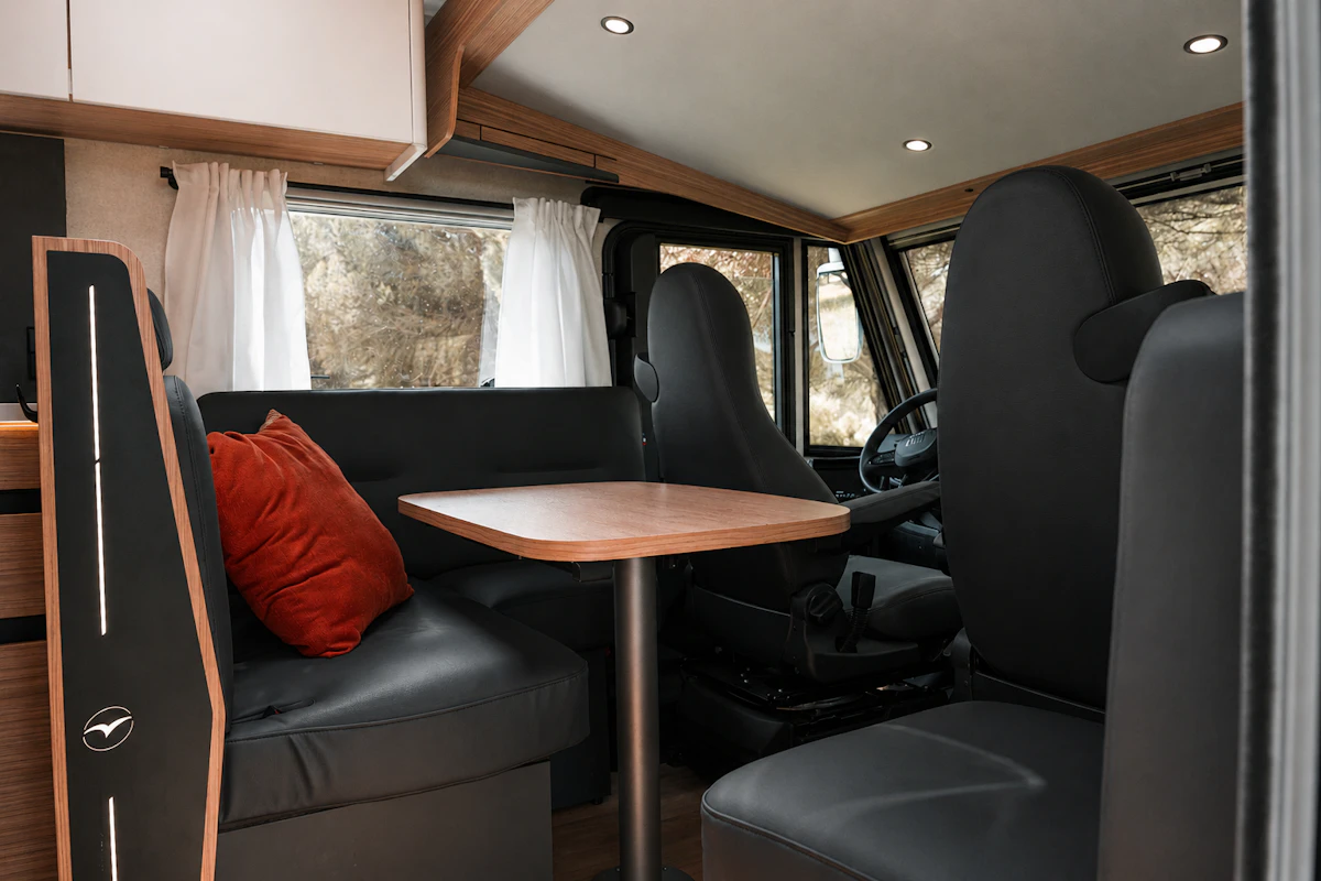 Van interior with front seats, dining table, side bench, overhead cabinets, and window curtains visible.