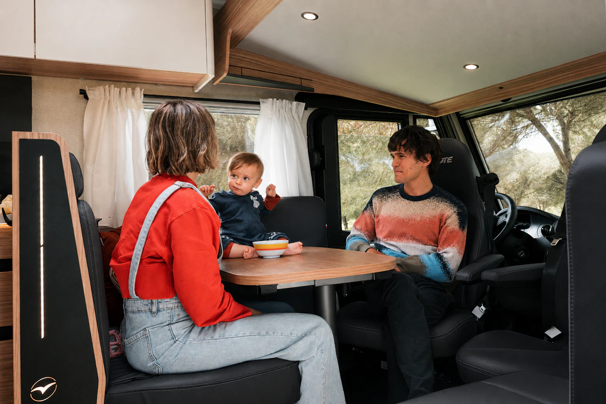 Van interior with dining table, swivel seats, upper storage cabinets, and curtained windows.
