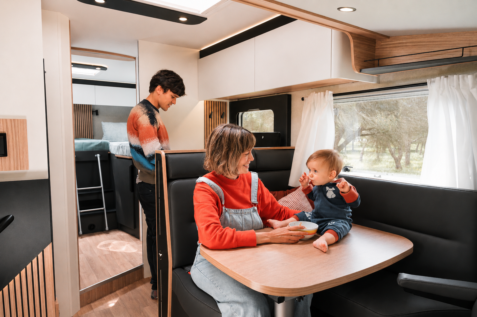 Van interior with dining table, black bench seats, overhead cabinets, large window with curtains, and a side kitchen area.